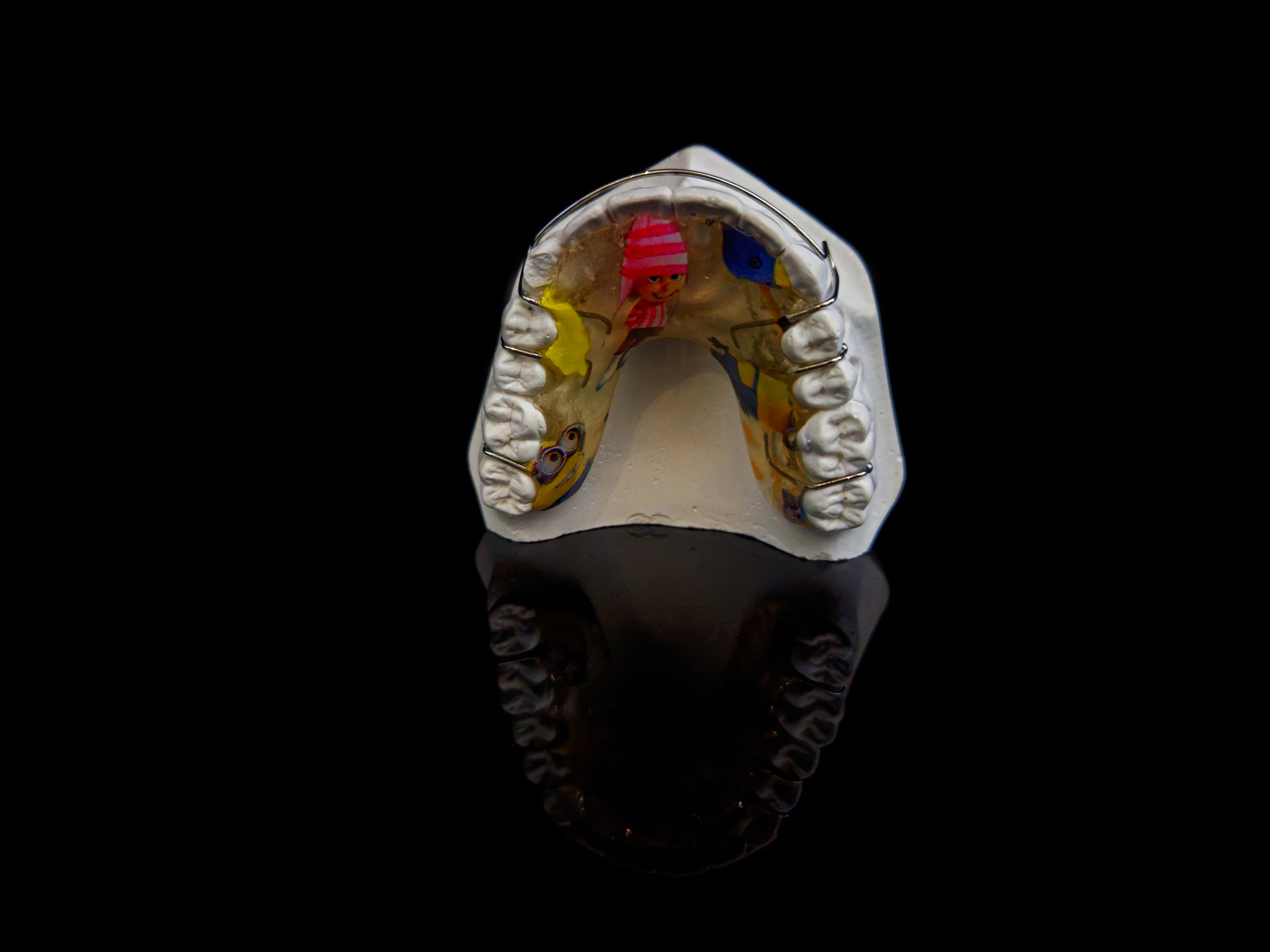 Model gipsowy szczęki z aparatem ortodontycznym ozdobionym kolorowymi postaciami z bajek, na czarnym, błyszczącym tle.