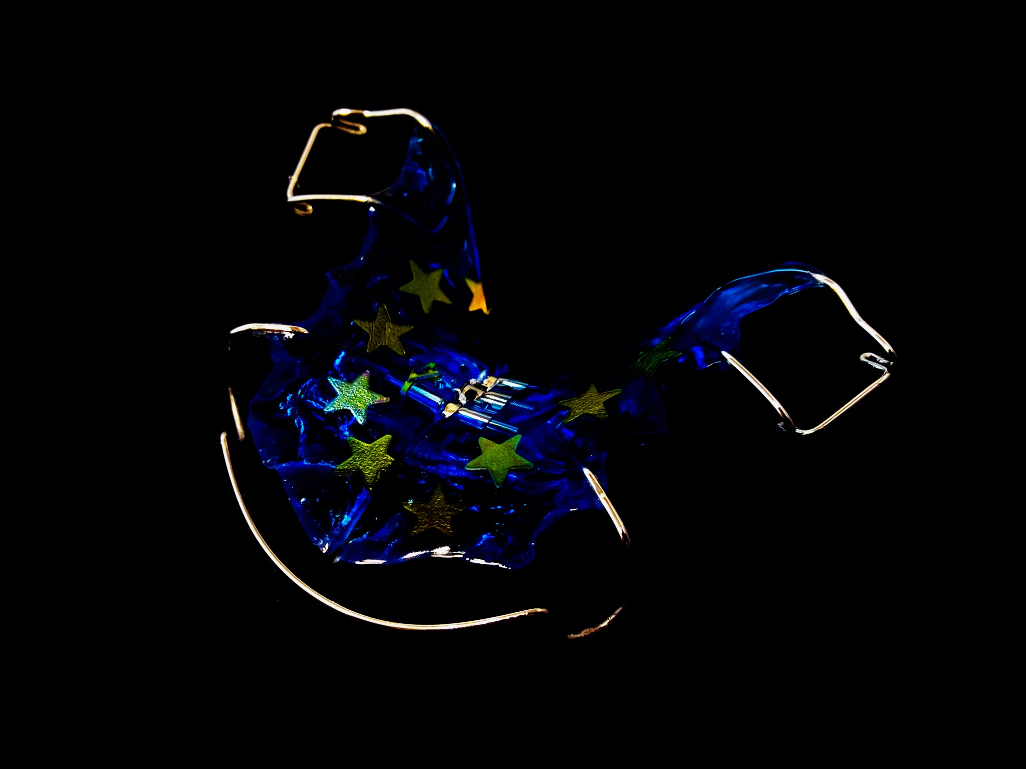 Aparat ortodontyczny ruchomy z niebieską płytką ozdobioną zielonymi i złotymi gwiazdkami, na czarnym tle.