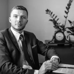 Portret mężczyzny w garniturze siedzącego przy biurku z lampą, zegarem i laptopem, czarno-białe zdjęcie.