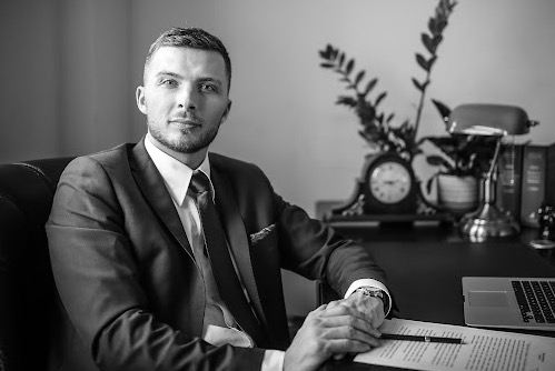 Portret mężczyzny w garniturze siedzącego przy biurku z lampą, zegarem i laptopem, czarno-białe zdjęcie.