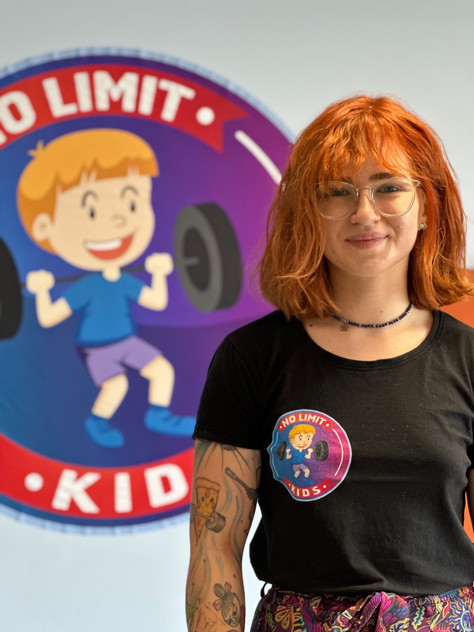 Uśmiechnięta kobieta w okularach i czarnej koszulce z logo 'No Limit Kids' na tle ściany z podobnym, większym logo przedstawiającym chłopca podnoszącego sztangę, widoczny tatuaż na ramieniu.