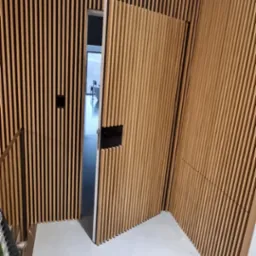 Nowoczesne drzwi drewniane z pionowymi listwami, częściowo otwarte, wkomponowane w ścianę z identycznymi listwami, tworzące spójny design wnętrza. Widoczna klamka w kolorze czarnym i fragment...