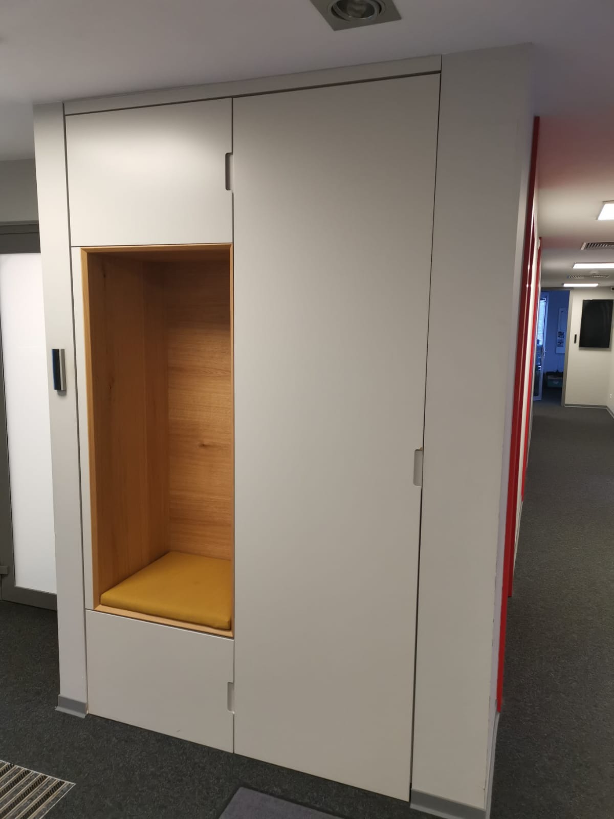 Biała szafa wnękowa z drewnianą wnęką do siedzenia z żółtą poduszką, w biurze.