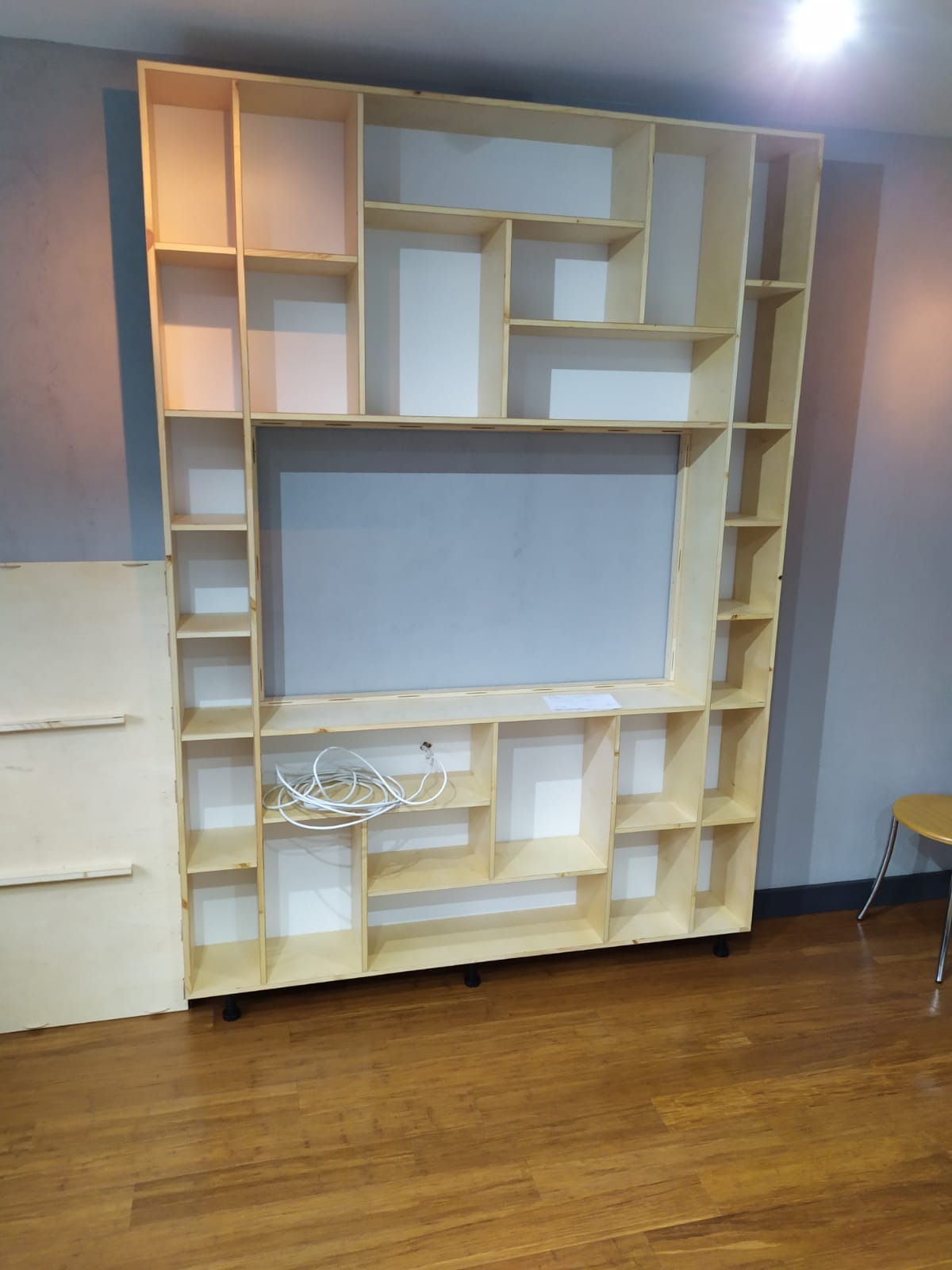 Konstrukcja regału z jasnego drewna z licznymi półkami, częściowo pomalowanymi na biało, na kółkach, z widocznymi kablami w dolnej części i dodatkowym panelem po lewej stronie, na tle drewnianej...