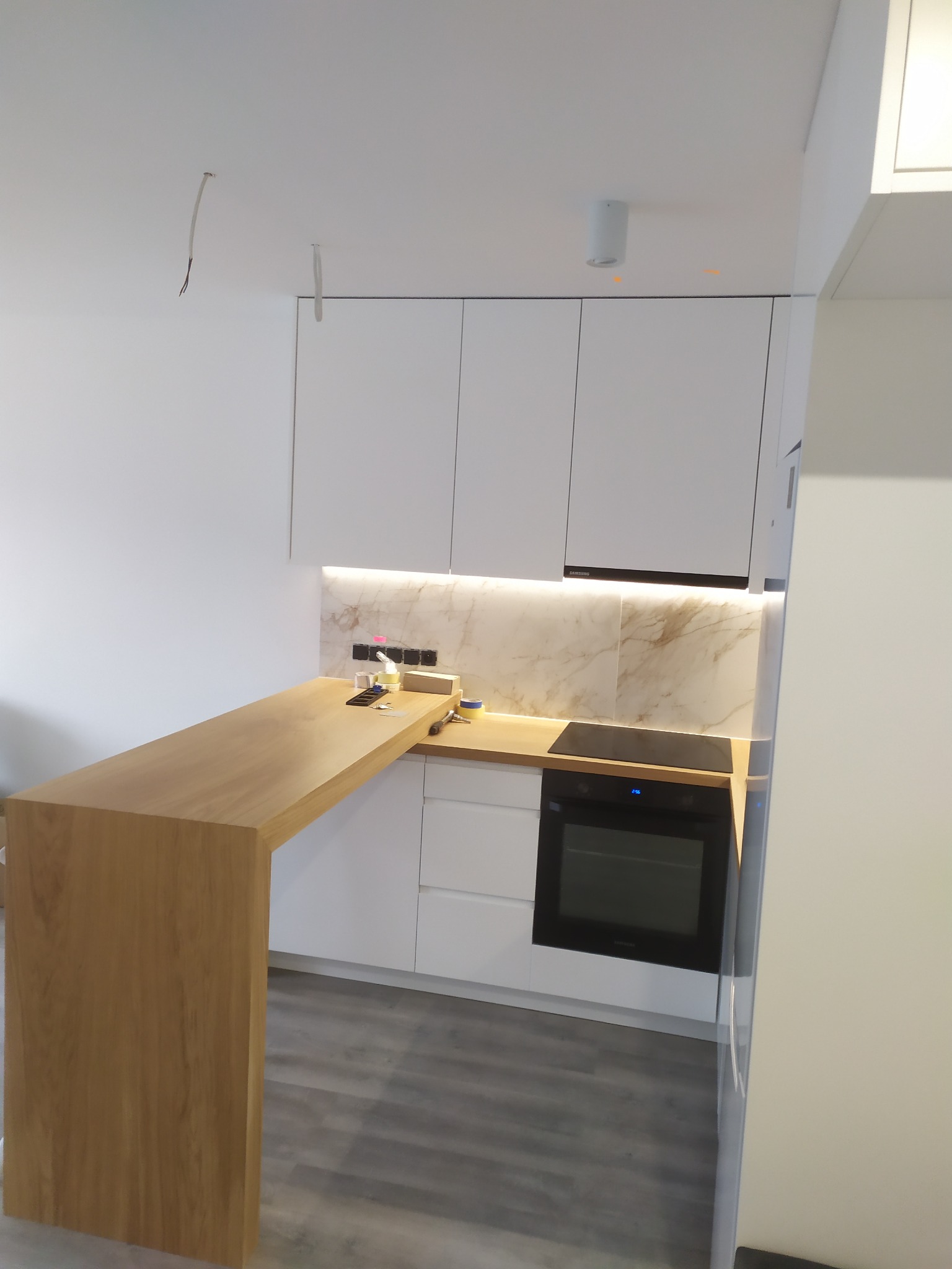 Nowoczesna kuchnia z białymi szafkami, drewnianym blatem roboczym i marmurowym panelem nad blatem, widoczne niedokończone prace elektryczne na ścianie.