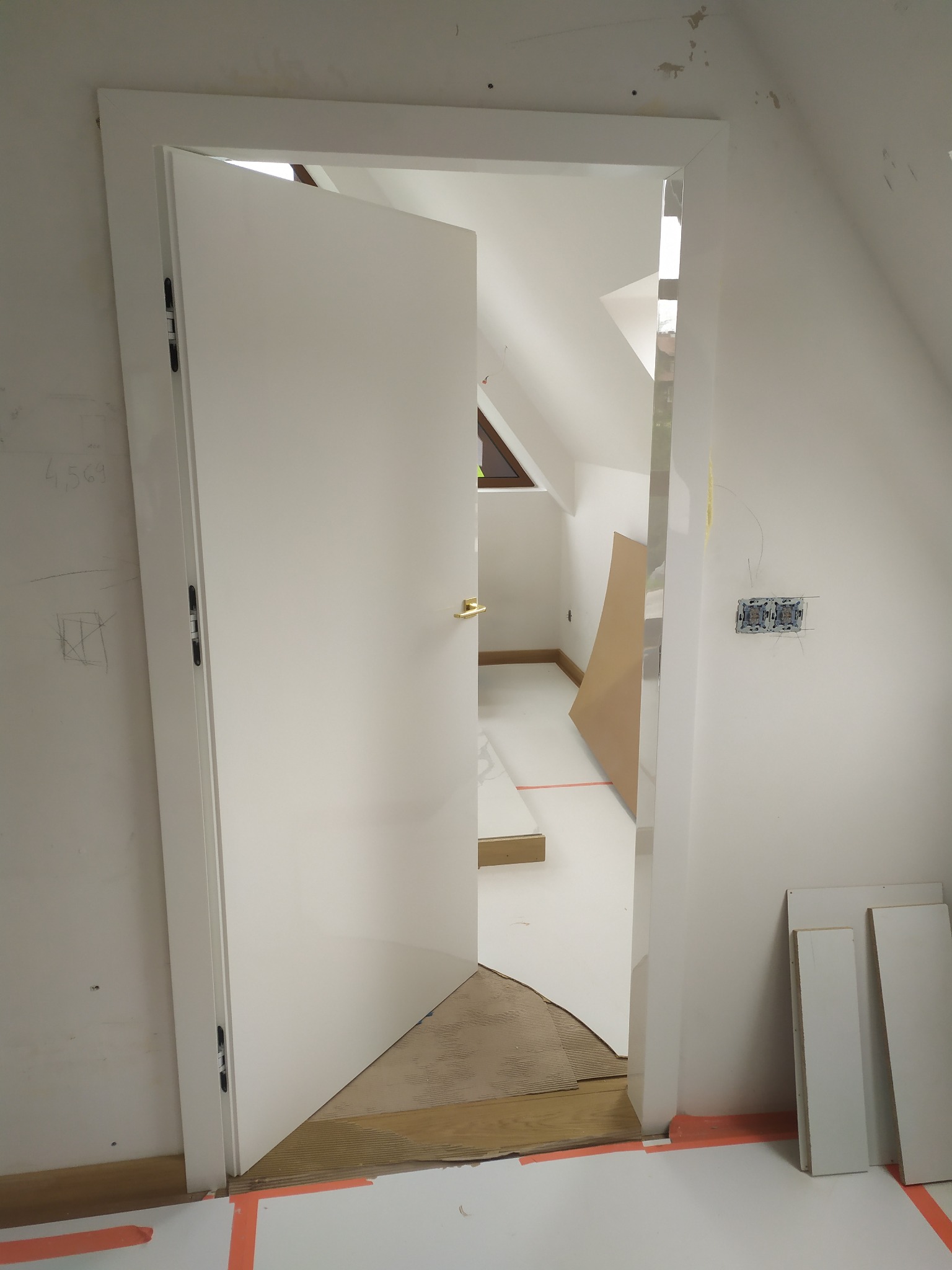 Montaż białych drzwi wewnętrznych z ościeżnicą w pokoju na poddaszu, widoczne niedokończone prace wykończeniowe, zabezpieczona podłoga, wystające kable elektryczne.