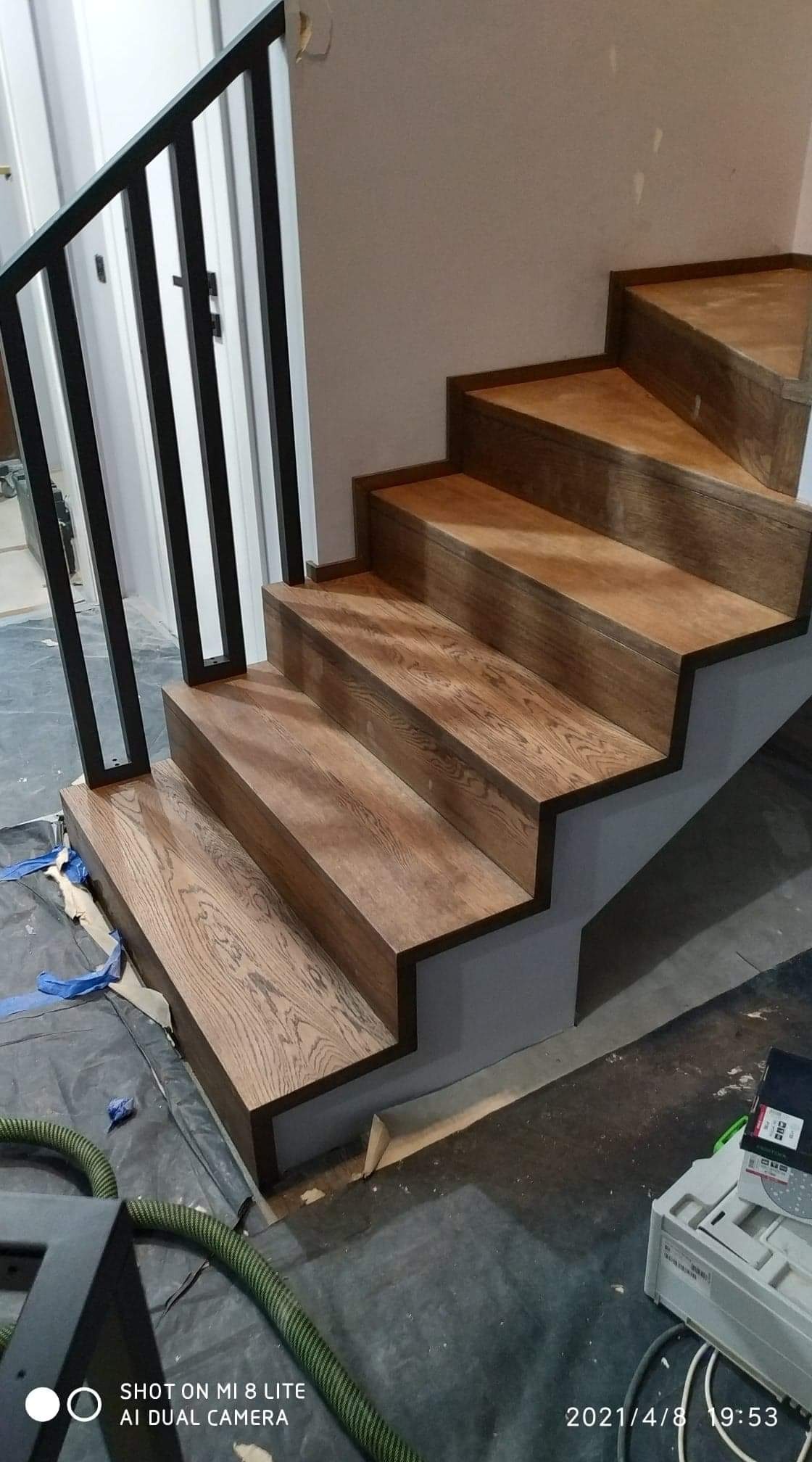 Schody drewniane o dębowym wykończeniu z czarną, metalową balustradą, widoczne w trakcie montażu, z folią ochronną i narzędziami na podłodze.