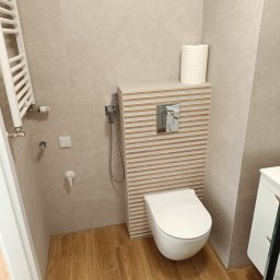 Bud Rud - Nowoczesna toaleta z podtynkową spłuczką, wykończona płytkami w odcieniach beżu i brązu. Na ścianie grzejnik drabinkowy i bidetka. Podłoga wyłożona płytkami imitującymi drewno.