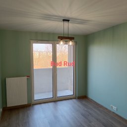 Bud Rud - Jasne wnętrze po remoncie: panele podłogowe, świeżo pomalowane ściany w odcieniach zieleni, nowoczesna lampa wisząca i balkon z widokiem na drzewa.