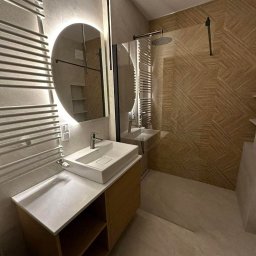 Bud Rud - Nowoczesna łazienka z owalnym lustrem podświetlanym LED, białą umywalką nablatową na drewnianej szafce i kabiną prysznicową z deszczownicą.