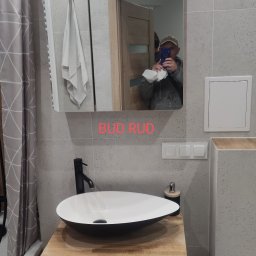 Bud Rud - Nowoczesna łazienka z umywalką nablatową na drewnianym blacie, szara płytka na ścianach, lustro z oświetleniem LED. Widoczne elementy armatury i akcesoria łazienkowe.