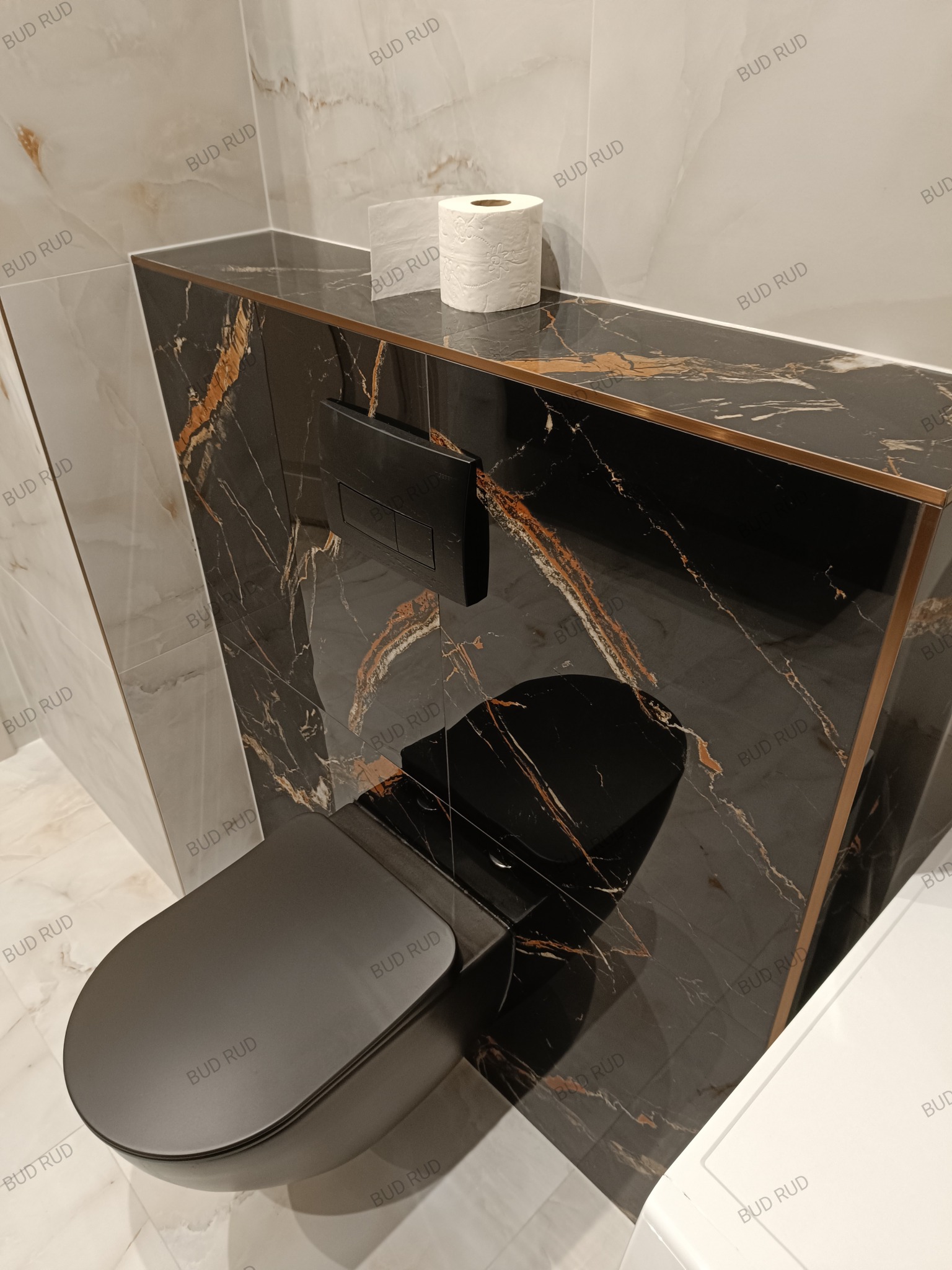 Nowoczesna toaleta w zabudowie z czarnego marmuru ze złotymi żyłami, z czarną miską WC i przyciskiem spłukującym. Na blacie rolka papieru toaletowego.