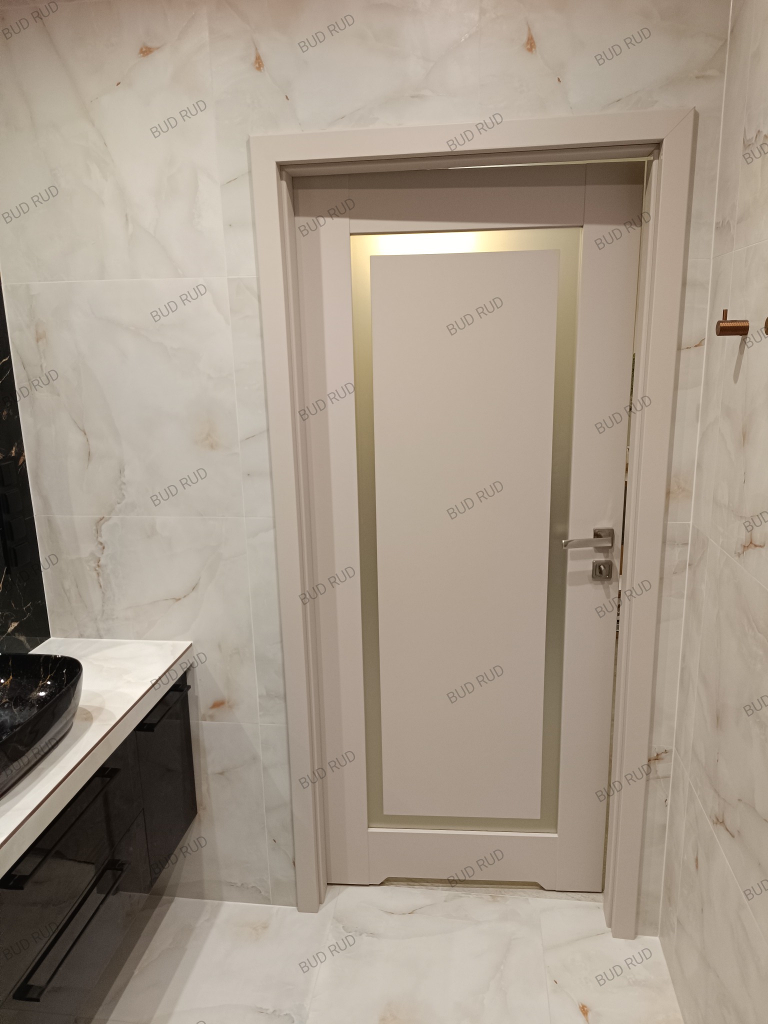 Zamknięte, kremowe drzwi z delikatnym frezem w nowoczesnej łazience z marmurowymi płytkami i czarną umywalką nablatową. Minimalistyczny design.