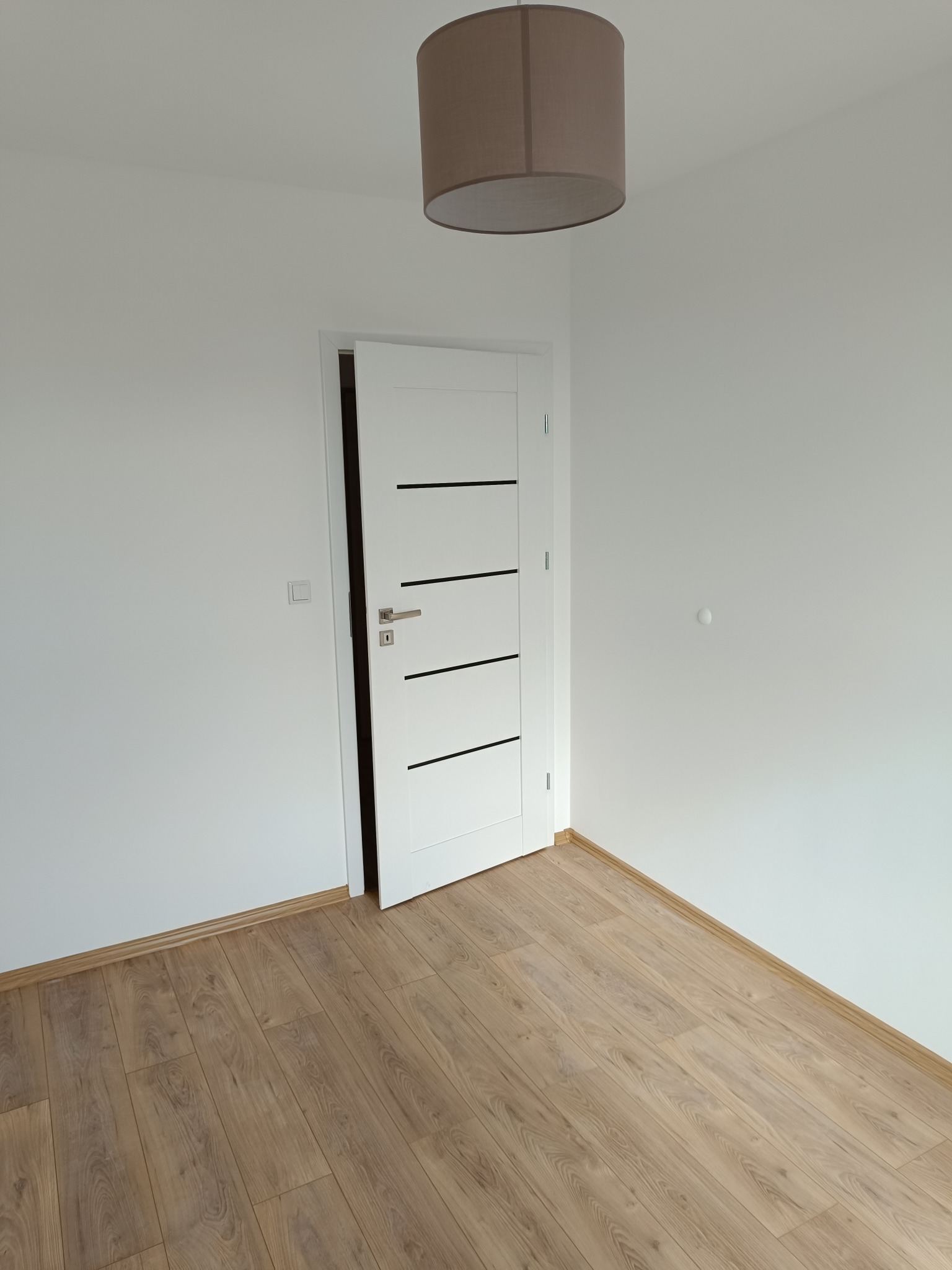 Jasne panele podłogowe w nowym pokoju z białą ścianą i otwartymi, nowoczesnymi drzwiami z poziomymi, czarnymi paskami. Brązowa lampa sufitowa.
