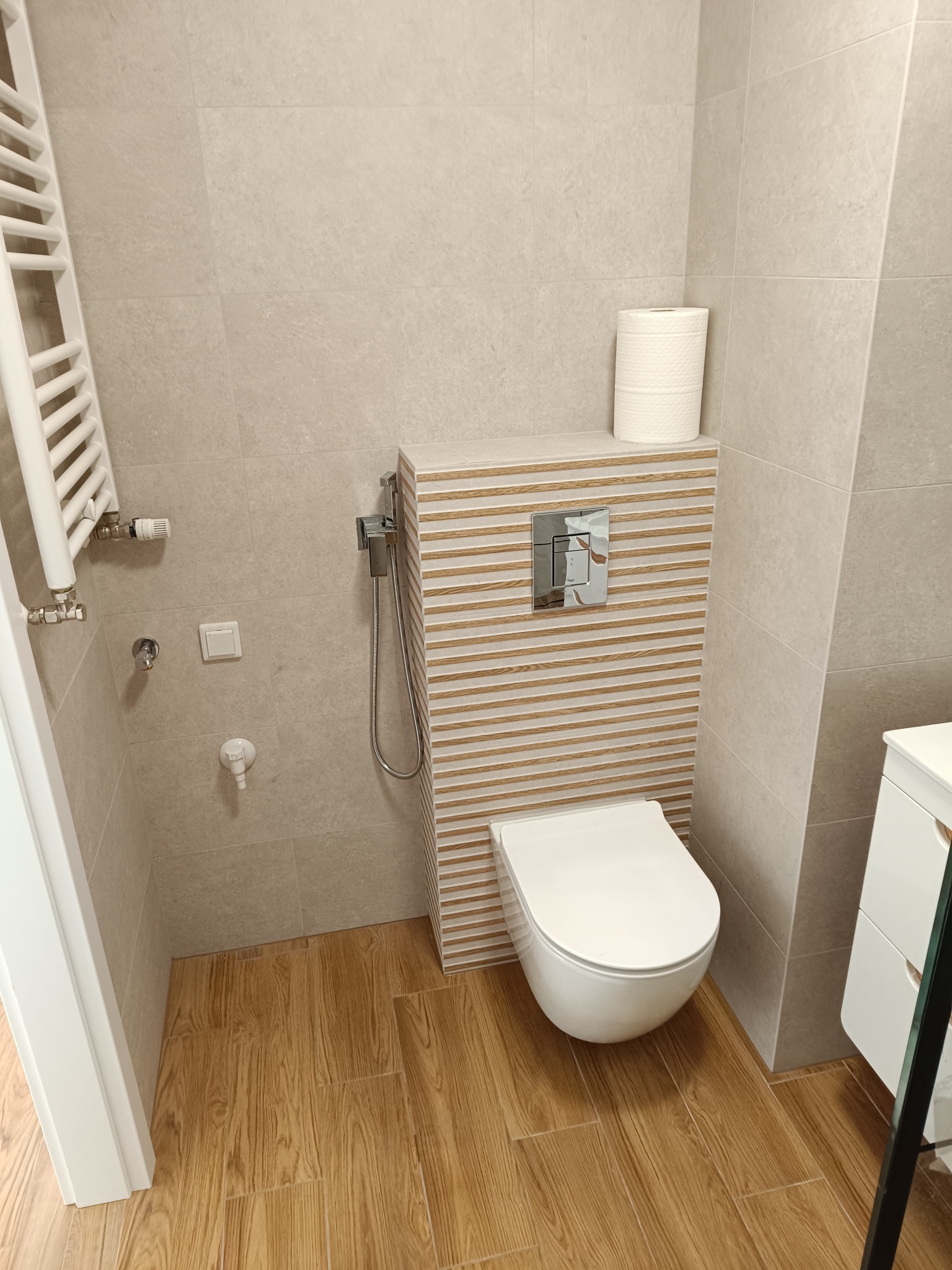Nowoczesna toaleta z podtynkową spłuczką, wykończona płytkami w odcieniach beżu i brązu. Na ścianie grzejnik drabinkowy i bidetka. Podłoga wyłożona płytkami imitującymi drewno.