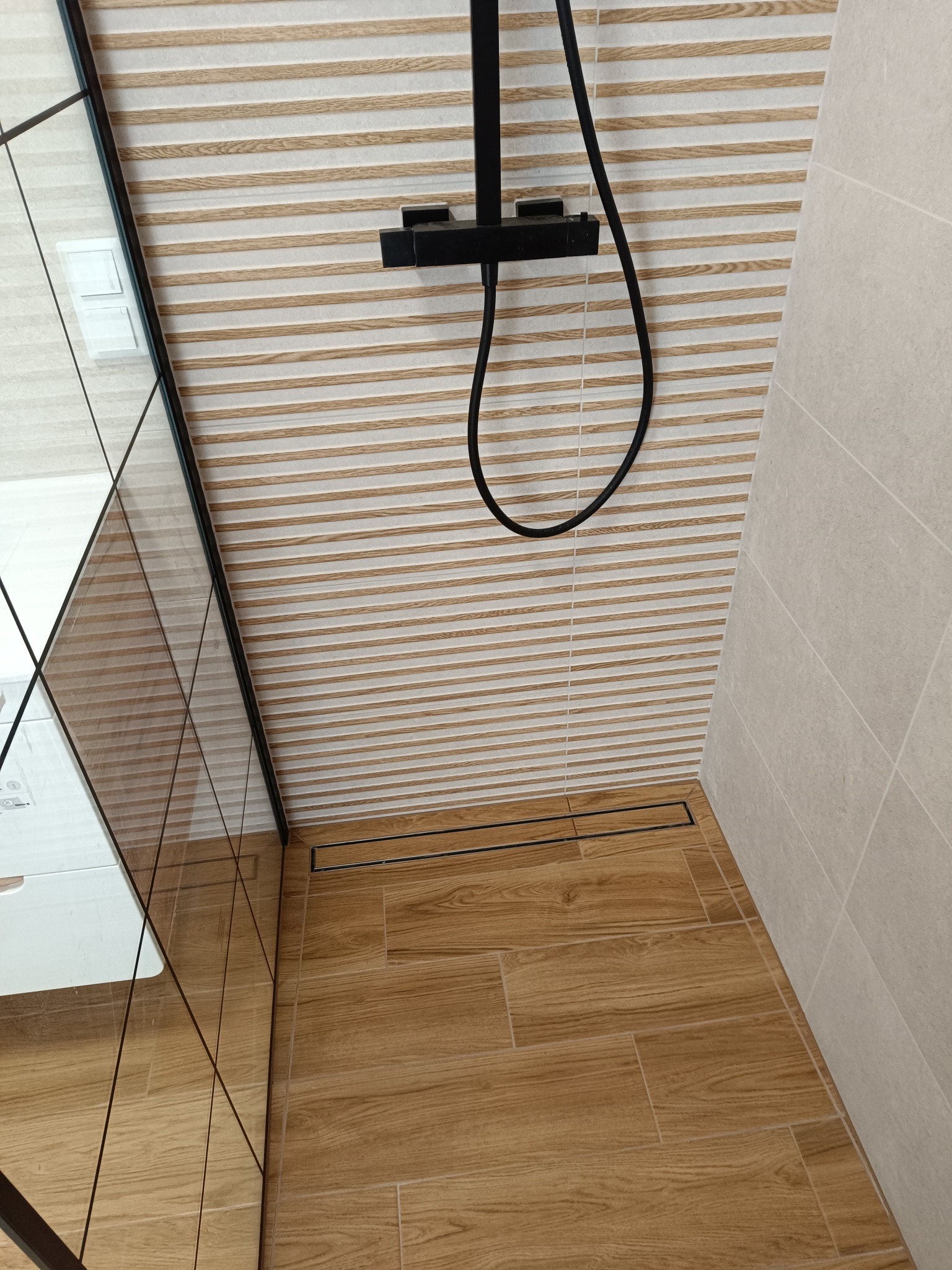 Nowoczesna kabina prysznicowa z drewnopodobnymi płytkami na podłodze i ścianie, czarna armatura, szklana ścianka. Minimalistyczny design z odpływem liniowym.