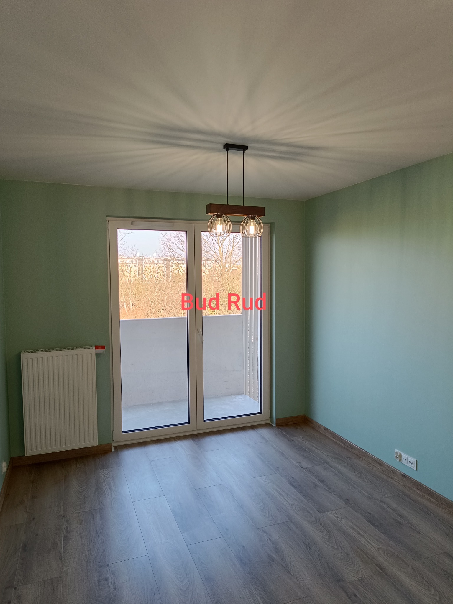 Jasne wnętrze po remoncie: panele podłogowe, świeżo pomalowane ściany w odcieniach zieleni, nowoczesna lampa wisząca i balkon z widokiem na drzewa.