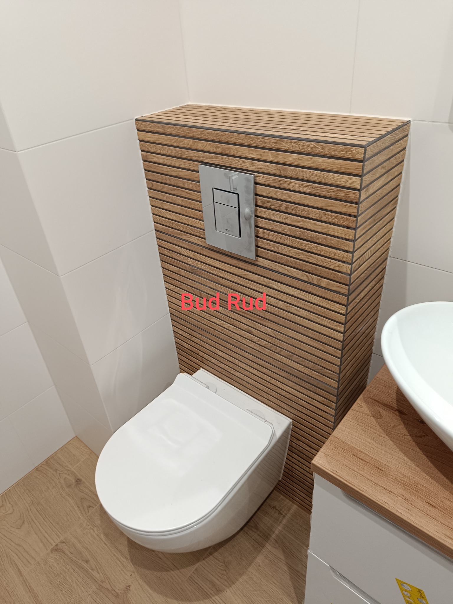 Nowoczesna toaleta podwieszana z obudową z drewnopodobnych paneli, chromowany przycisk spłukiwania, białe płytki i umywalka nablatowa. Wykończenie łazienki.