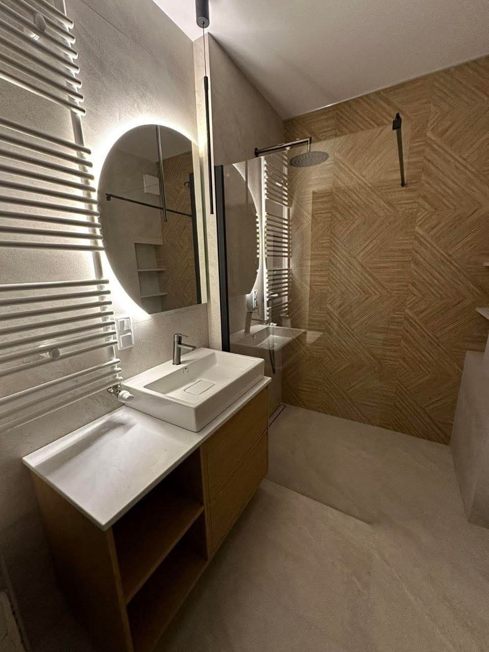 Nowoczesna łazienka z owalnym lustrem podświetlanym LED, białą umywalką nablatową na drewnianej szafce i kabiną prysznicową z deszczownicą.