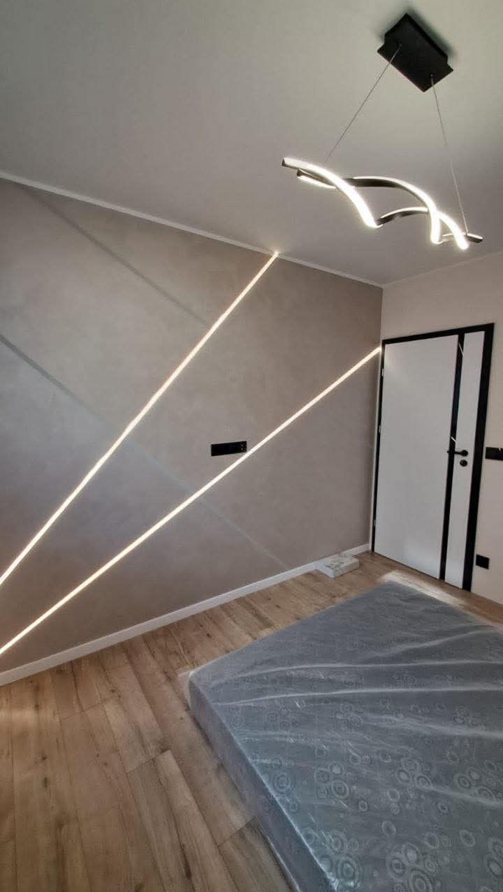 Pokój po remoncie: gładkie, beżowe ściany z dekoracyjnym oświetleniem LED wzdłuż skosów, drewniana podłoga i nowoczesna lampa sufitowa. Widoczne drzwi z czarnymi akcentami.