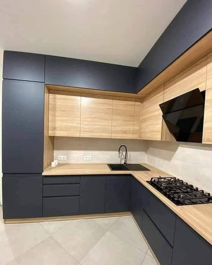 Nowoczesna kuchnia z ciemnymi, matowymi szafkami dolnymi i drewnopodobnymi górnymi. Czarny okap i zlewozmywak, jasny blat i płytki podłogowe. Minimalistyczny design.