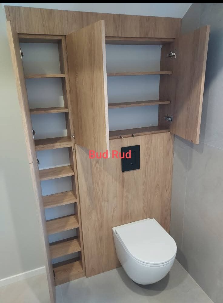 Zabudowa toalety z szafkami z drewna w jasnym odcieniu, z otwartymi drzwiczkami ukazującymi półki. Nowoczesna toaleta podwieszana, szara ściana w tle.