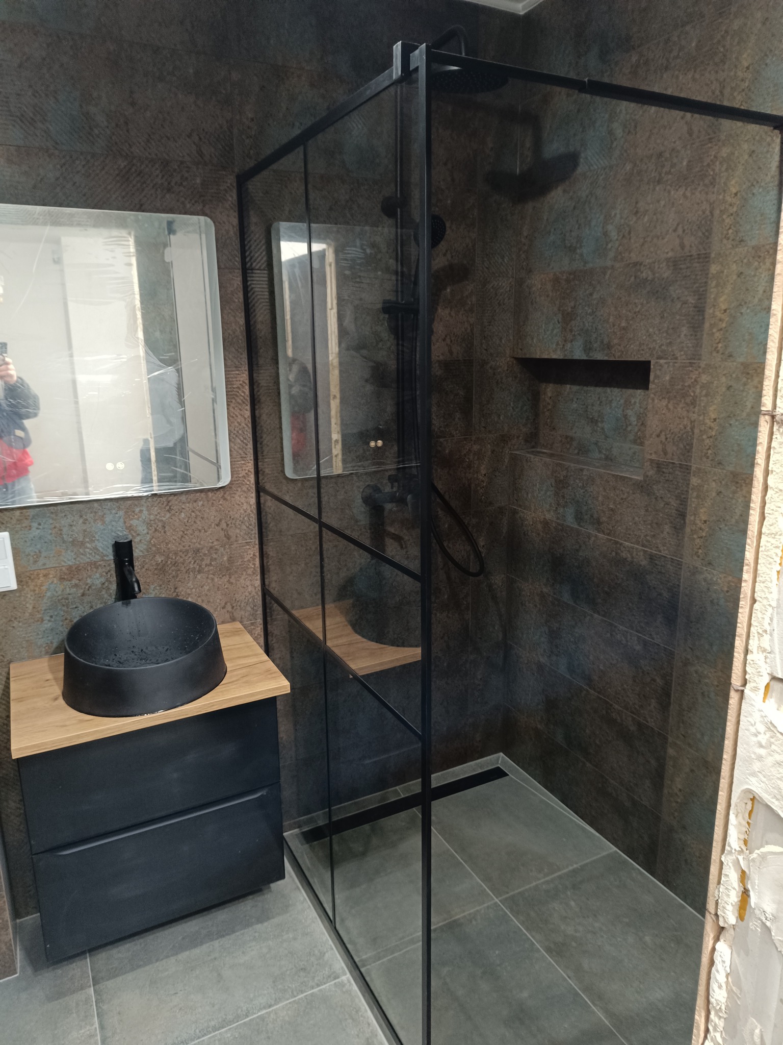 Nowoczesna łazienka z kabiną prysznicową typu walk-in, czarną armaturą i umywalką nablatową. Widoczne płytki imitujące kamień i lustro z podświetleniem LED.