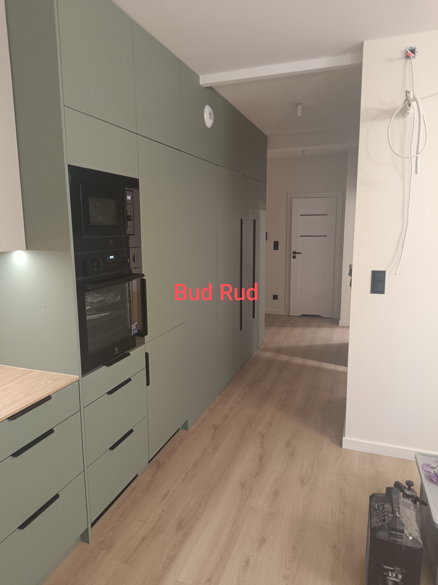 Nowoczesna kuchnia w odcieniach zieleni z zabudową meblową pod sufit, sprzęt AGD w zabudowie, panele podłogowe. Widok na korytarz z białą stolarką drzwiową.