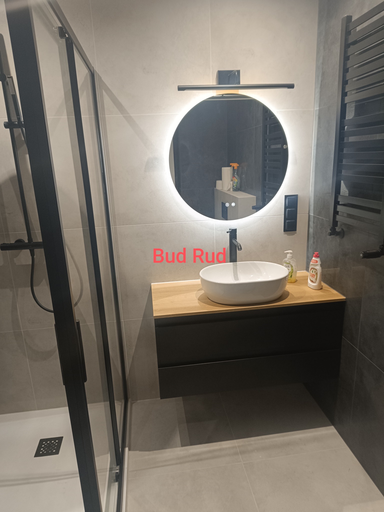 Nowoczesna łazienka z kabiną prysznicową, okrągłym lustrem z podświetleniem LED i umywalką nablatową na drewnianym blacie. Minimalistyczny design w szarościach.