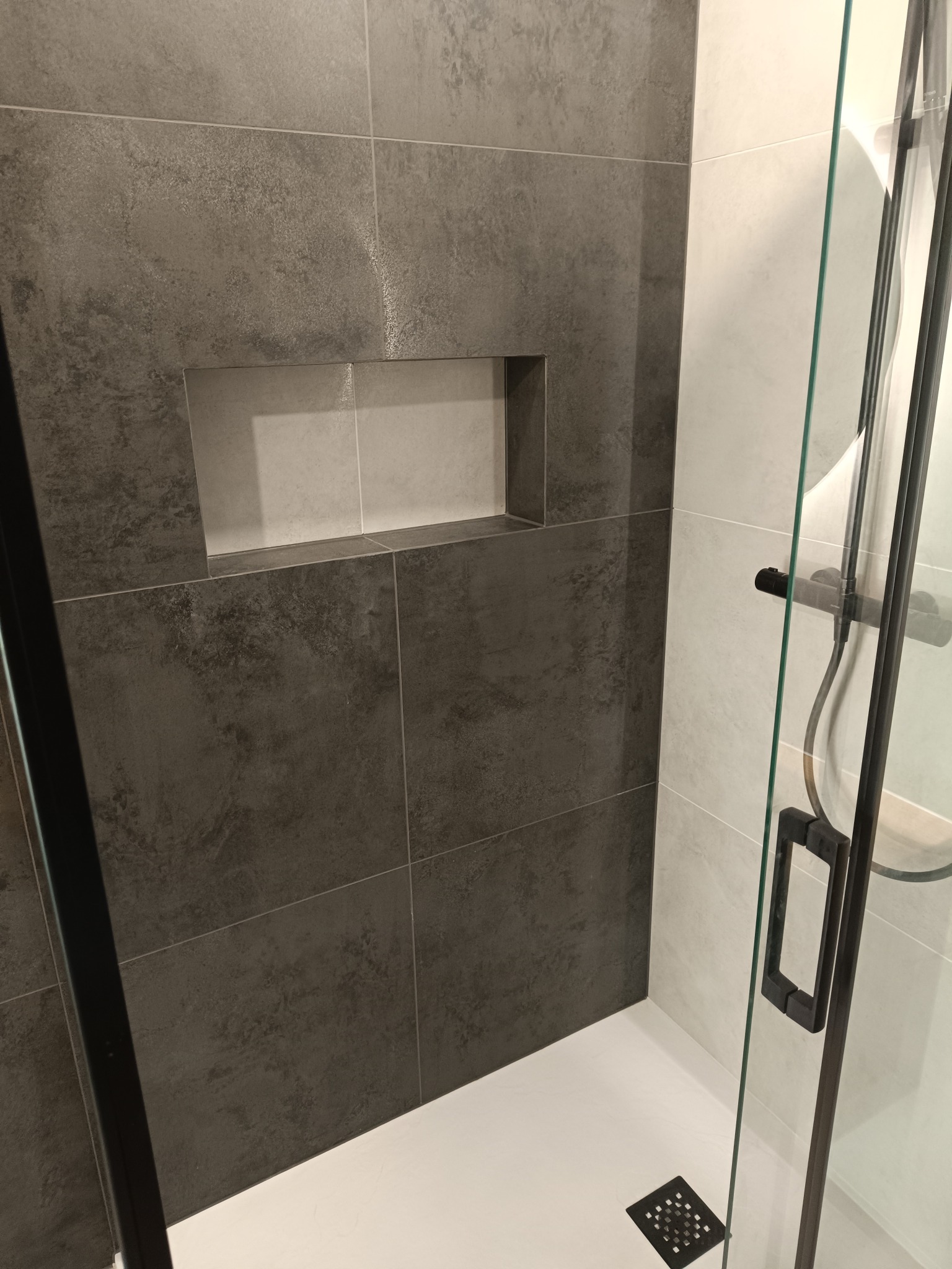 Nowoczesna kabina prysznicowa z szarymi płytkami imitującymi beton i wnęką w ścianie. Biały brodzik i czarne detale kabiny.