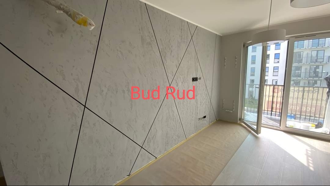 Ściana z dekoracyjną szpachlą w odcieniach szarości, z czarnymi, ukośnymi liniami tworzącymi geometryczny wzór. Widoczne okno balkonowe i fragment podłogi.