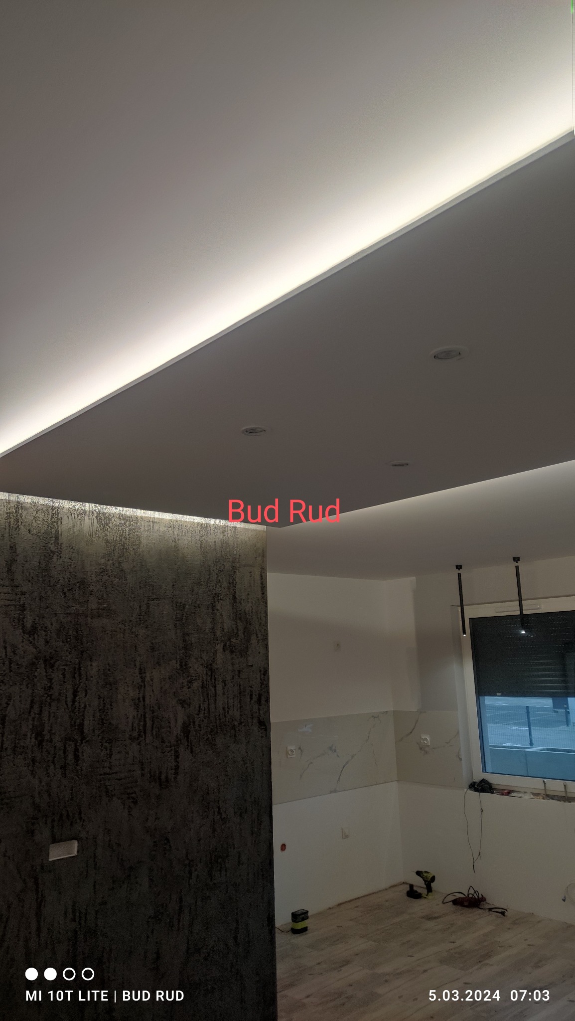 Nowoczesny sufit podwieszany z oświetleniem LED wzdłuż krawędzi, widoczne halogeny, ściana z dekoracyjną fakturą, fragment okna i podłogi w jasnym wnętrzu.