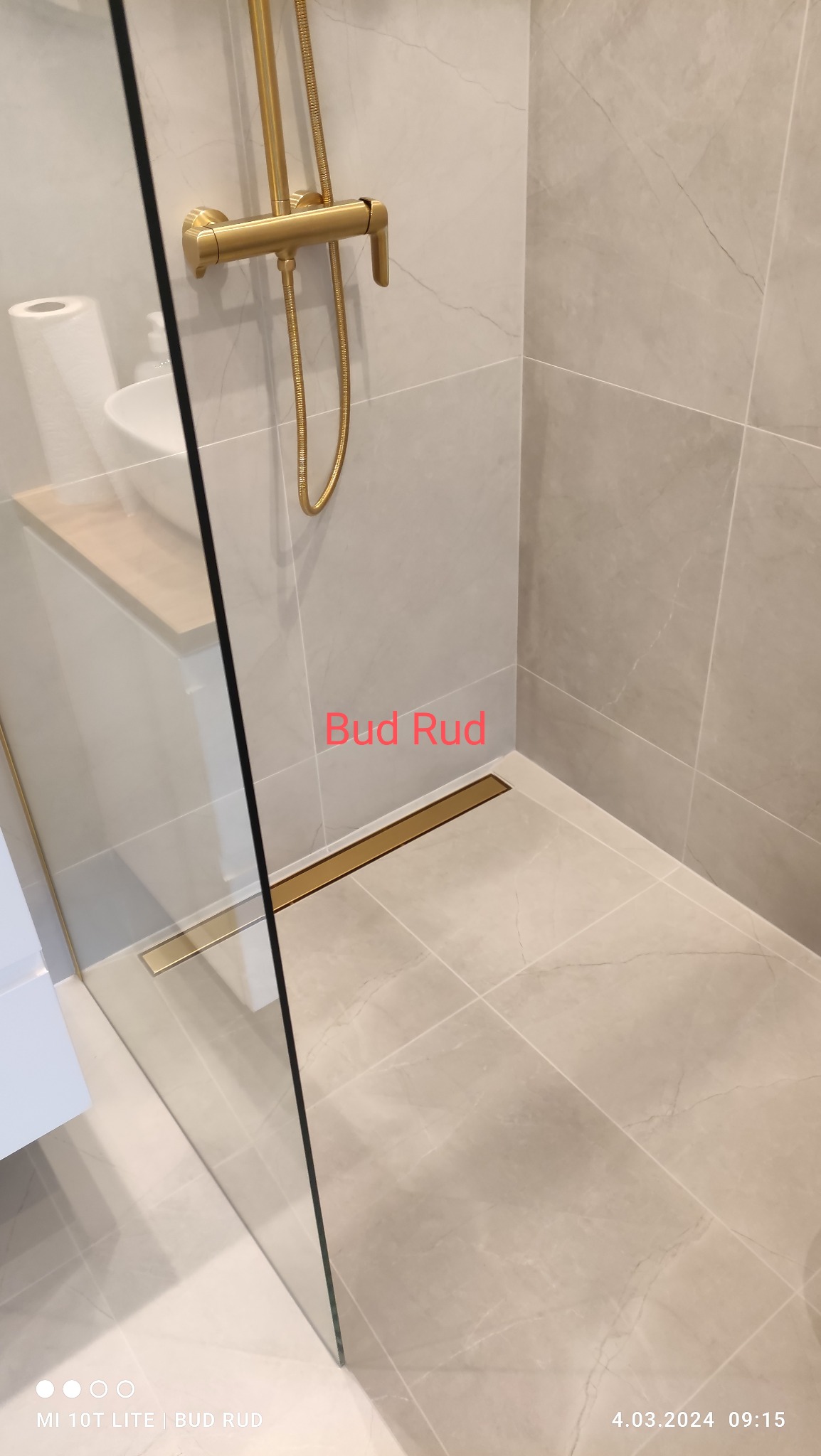 Nowoczesna kabina prysznicowa z odpływem liniowym w kolorze złotym, wykończona płytkami imitującymi marmur. Widoczna armatura w odcieniach złota i szklana ściana kabiny.