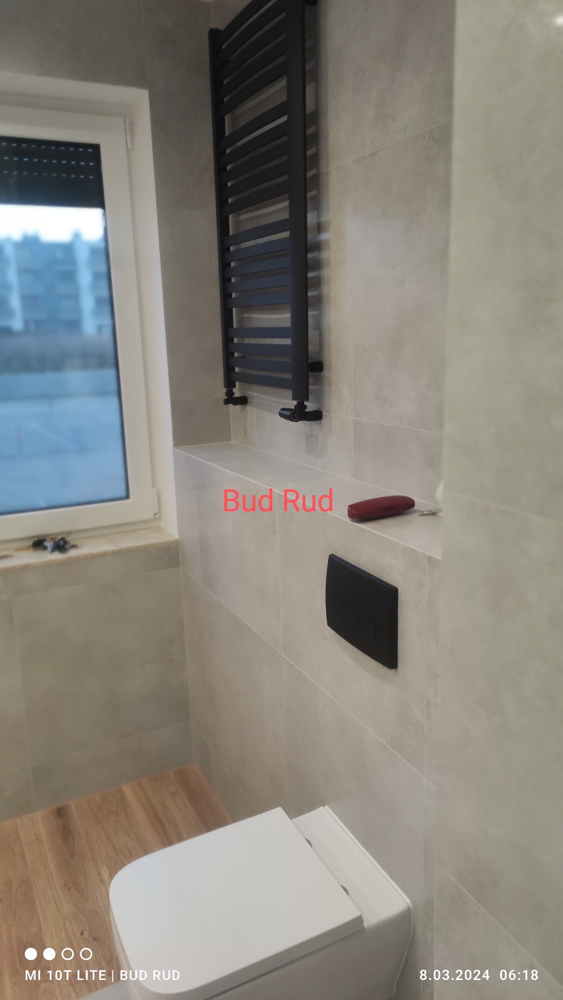 Wykończona łazienka z beżowymi płytkami, wiszącą toaletą i czarnym grzejnikiem drabinkowym. Widoczne detale: przycisk spłuczki, drewnopodobna podłoga i okno z roletą.