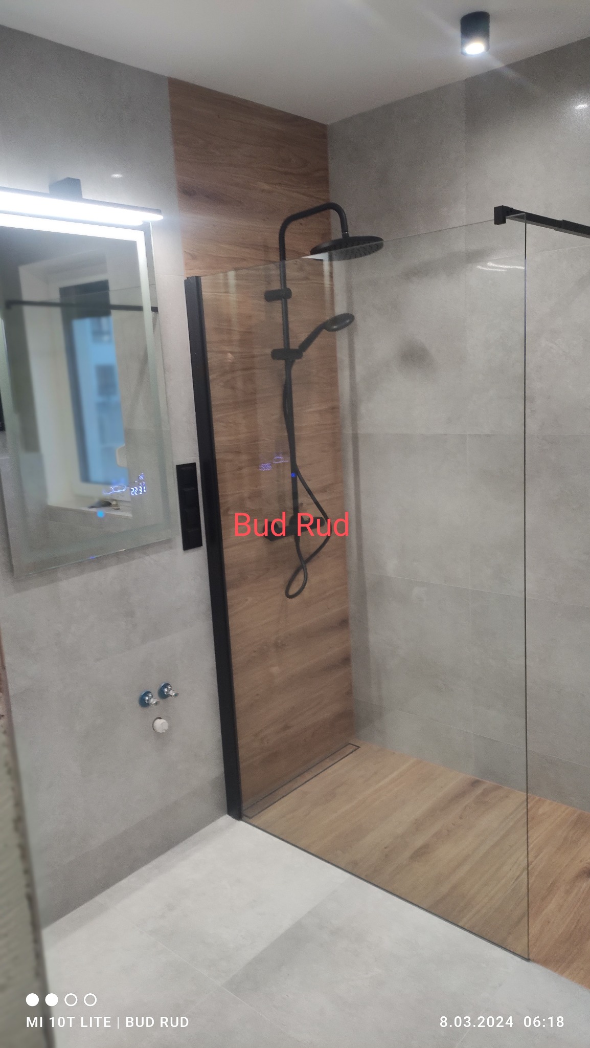 Nowoczesna kabina prysznicowa walk-in z czarną armaturą i drewnopodobnymi płytkami w strefie prysznica. Szklana ściana z czarnymi profilami. Lustro z podświetleniem.