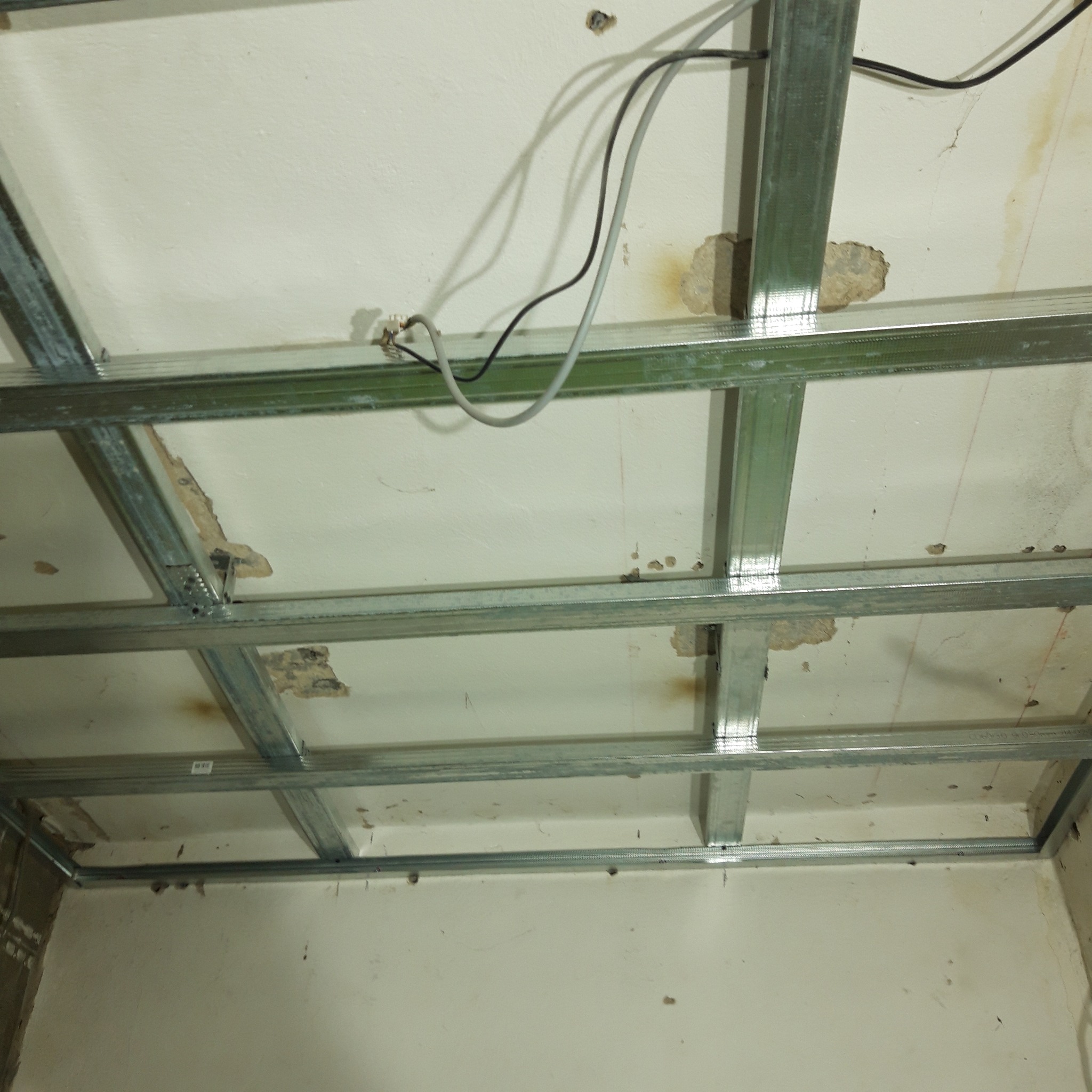 Metalowa konstrukcja sufitu podwieszanego z widocznymi przewodami elektrycznymi i fragmentami starego tynku na suficie, przygotowana do montażu płyt.