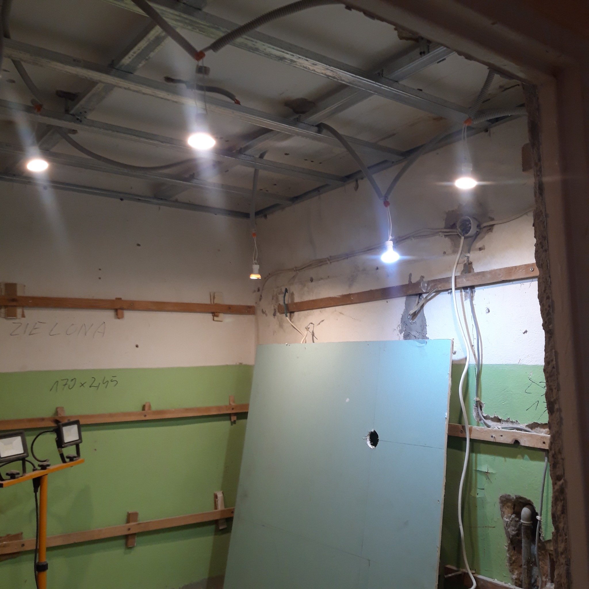 Wnętrze w trakcie prac przygotowawczych do montażu sufitu podwieszanego z płyt kartonowo-gipsowych, widoczne profile sufitowe, instalacja elektryczna, częściowo otynkowana ściana z zieloną farbą...