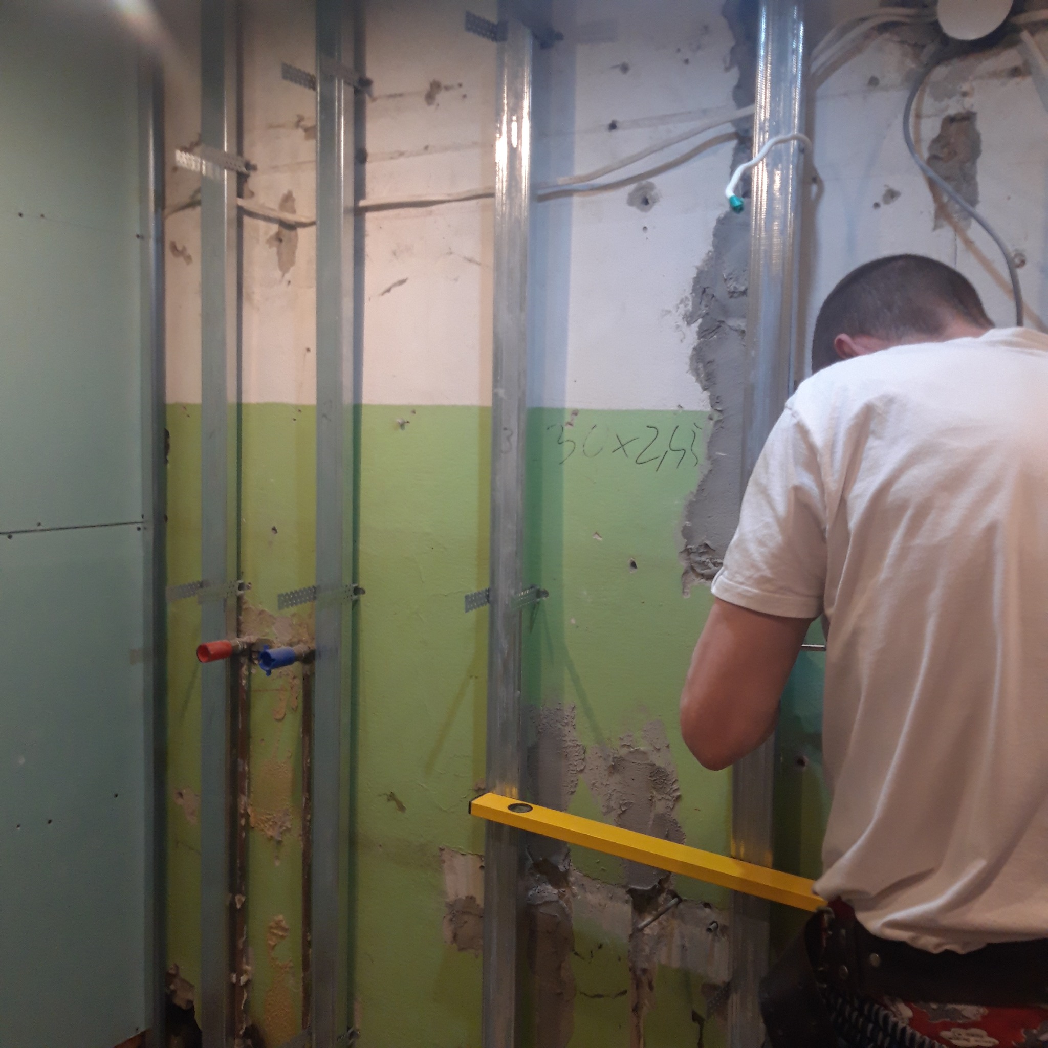 Instalacja metalowych profili na nierównej, zielonej ścianie z widocznymi śladami po instalacjach, poziomica w użyciu, przygotowanie pod płyty karton-gips.