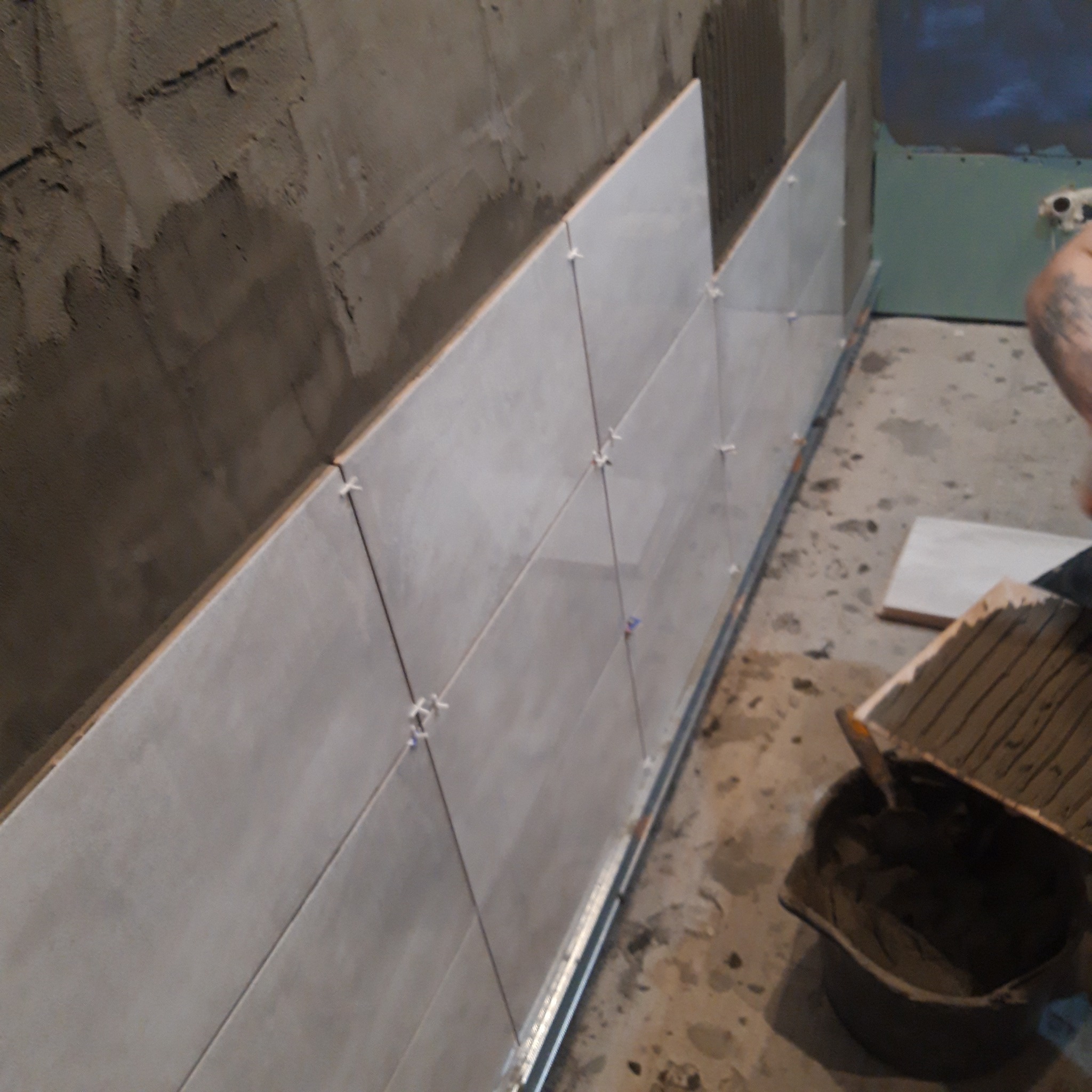 Ściana wyłożona jasnymi płytkami ceramicznymi, z krzyżykami dystansowymi, w trakcie prac wykończeniowych, widoczny fragment ściany z surowym tynkiem i wiadro z zaprawą.