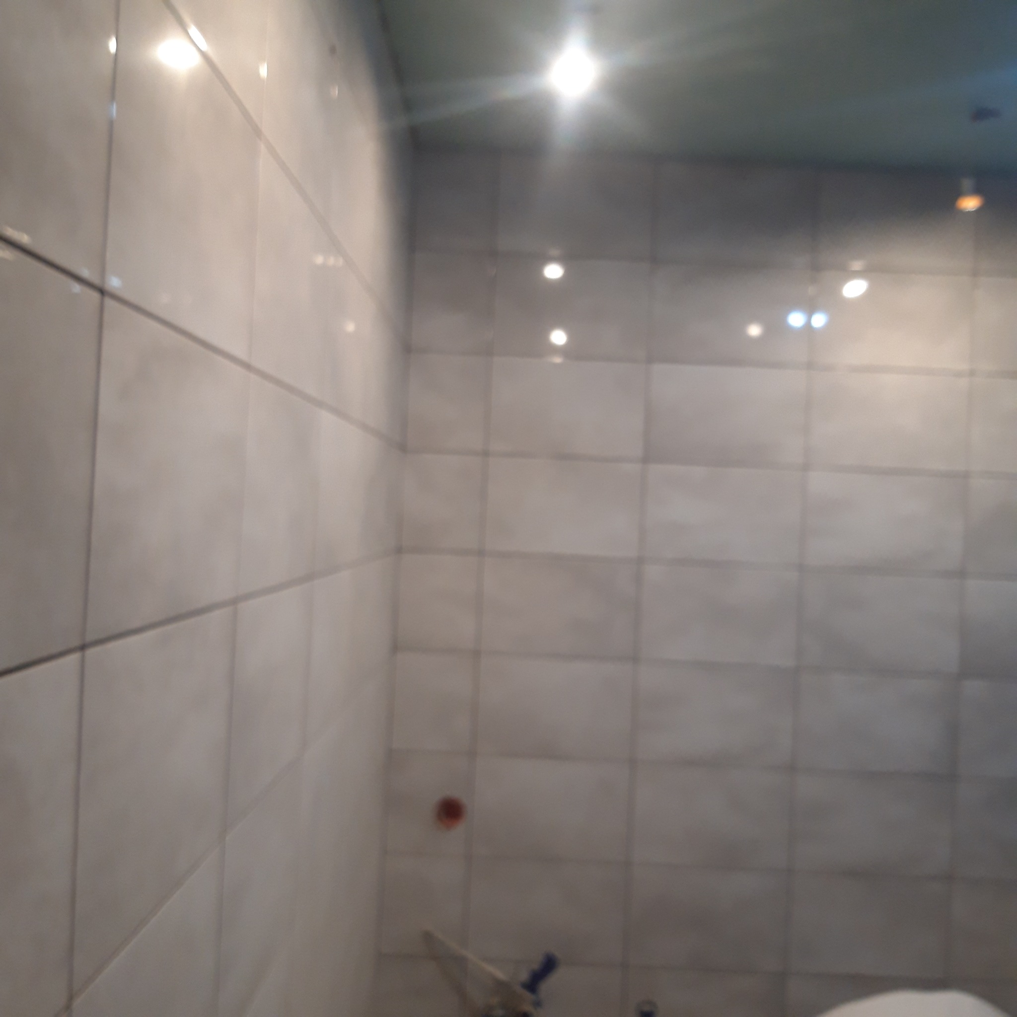 Ściana łazienki wyłożona szarymi płytkami ceramicznymi, widoczne fugi i odbicia światła od oświetlenia sufitowego.