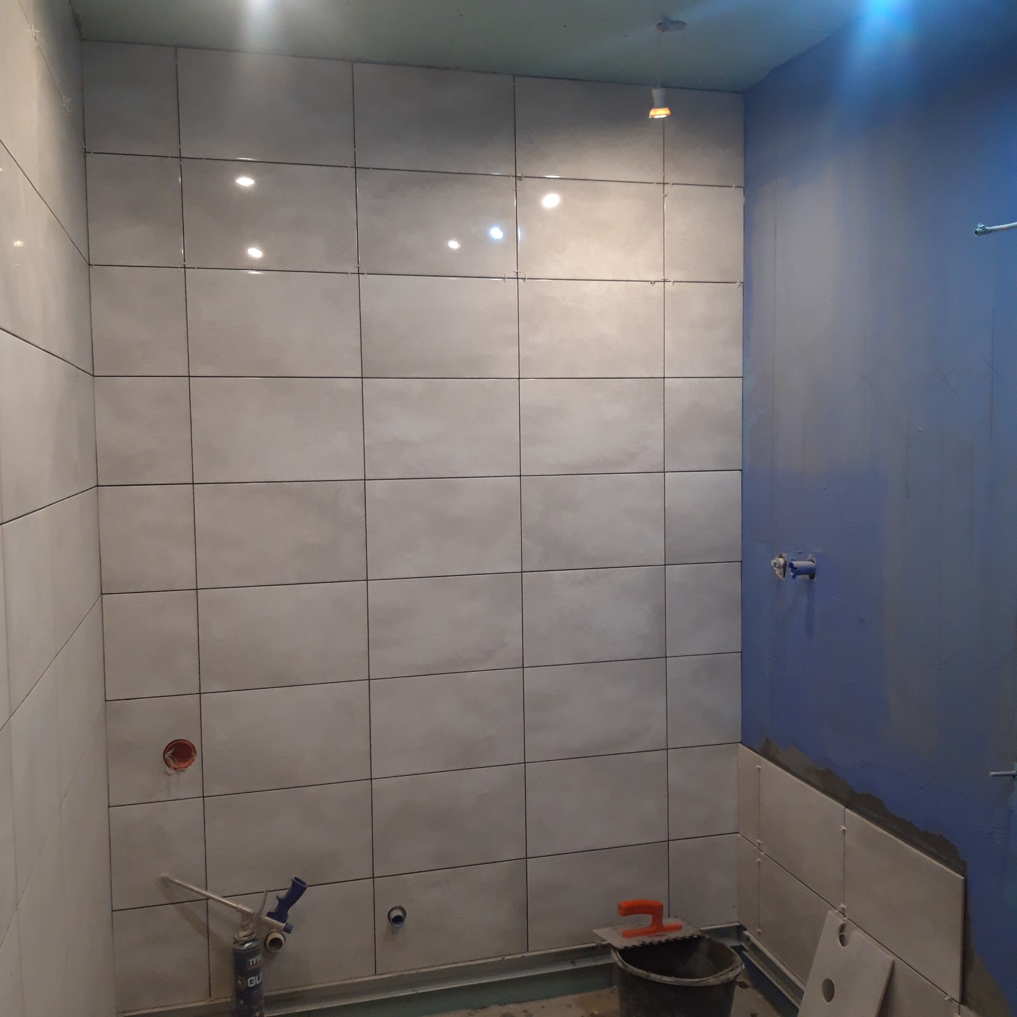 Ściana łazienki w trakcie kładzenia jasnych, kwadratowych płytek ceramicznych, widoczne narzędzia glazurnicze i niebieska ściana bez płytek.