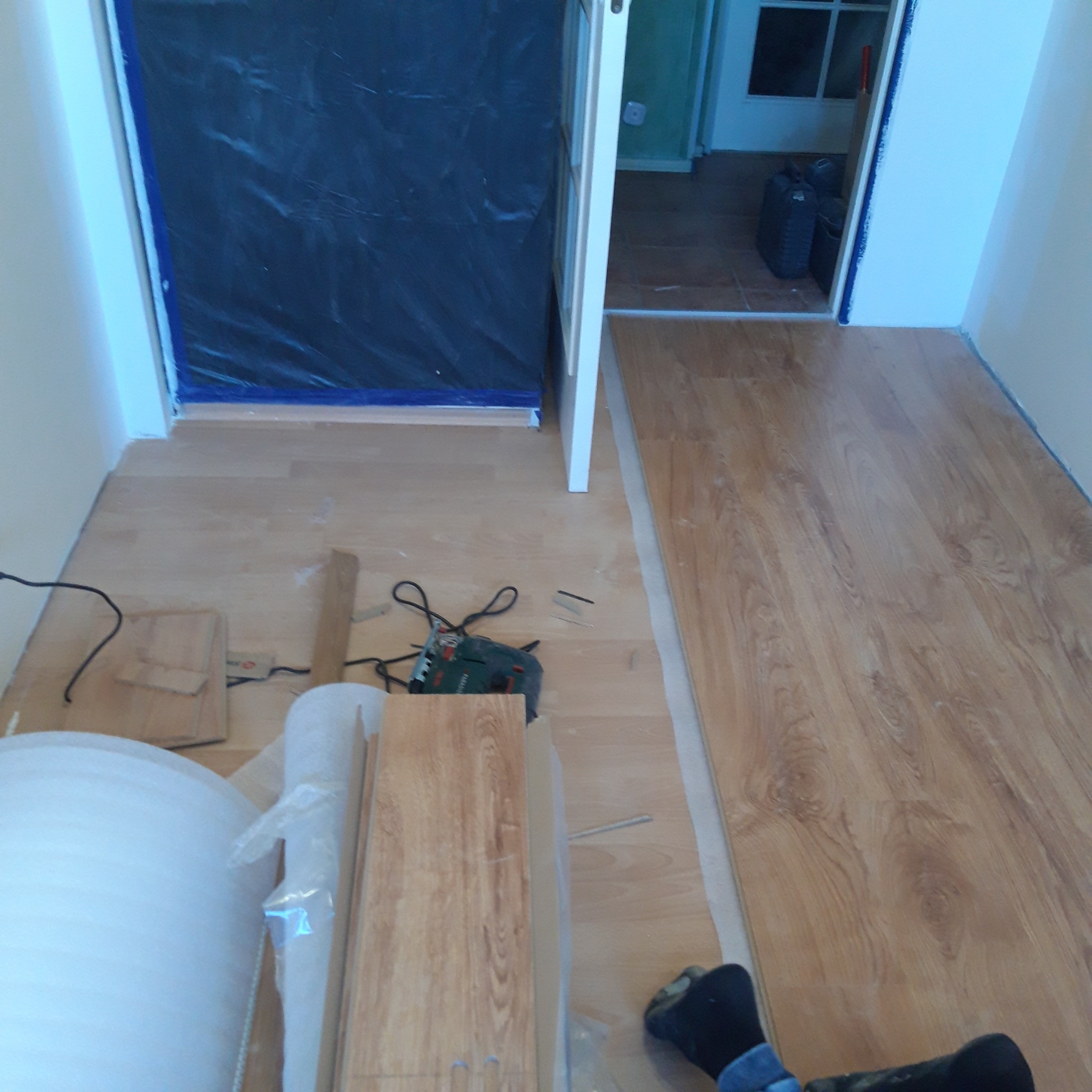 Proces układania paneli podłogowych w pomieszczeniu, widoczna część ułożonej podłogi, narzędzia, rolka pianki pod panele i zabezpieczona folią strefa wejścia.