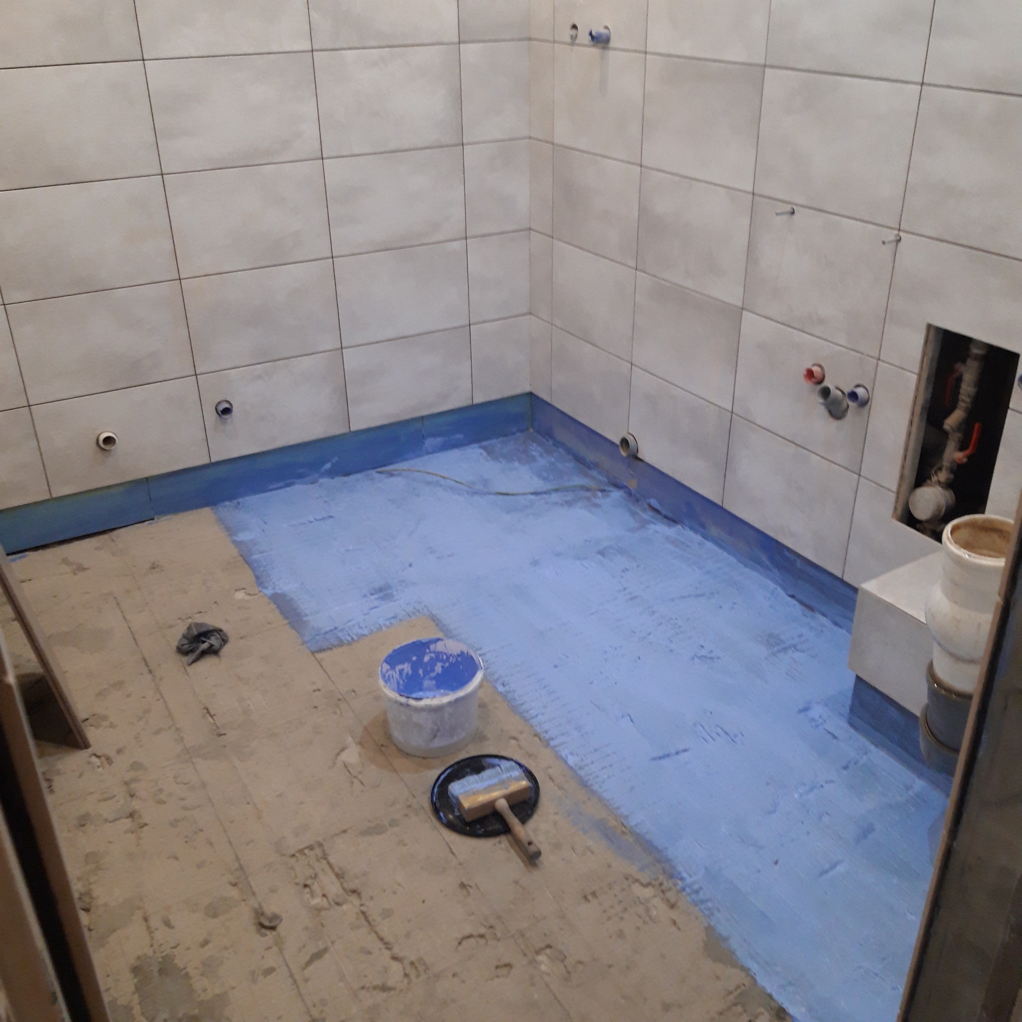 Przygotowanie do położenia płytek w łazience: podłoga pokryta niebieską hydroizolacją, widoczny pędzel do rozprowadzania, ściany wyłożone jasnymi płytkami, instalacja wodno-kanalizacyjna w ścianie.