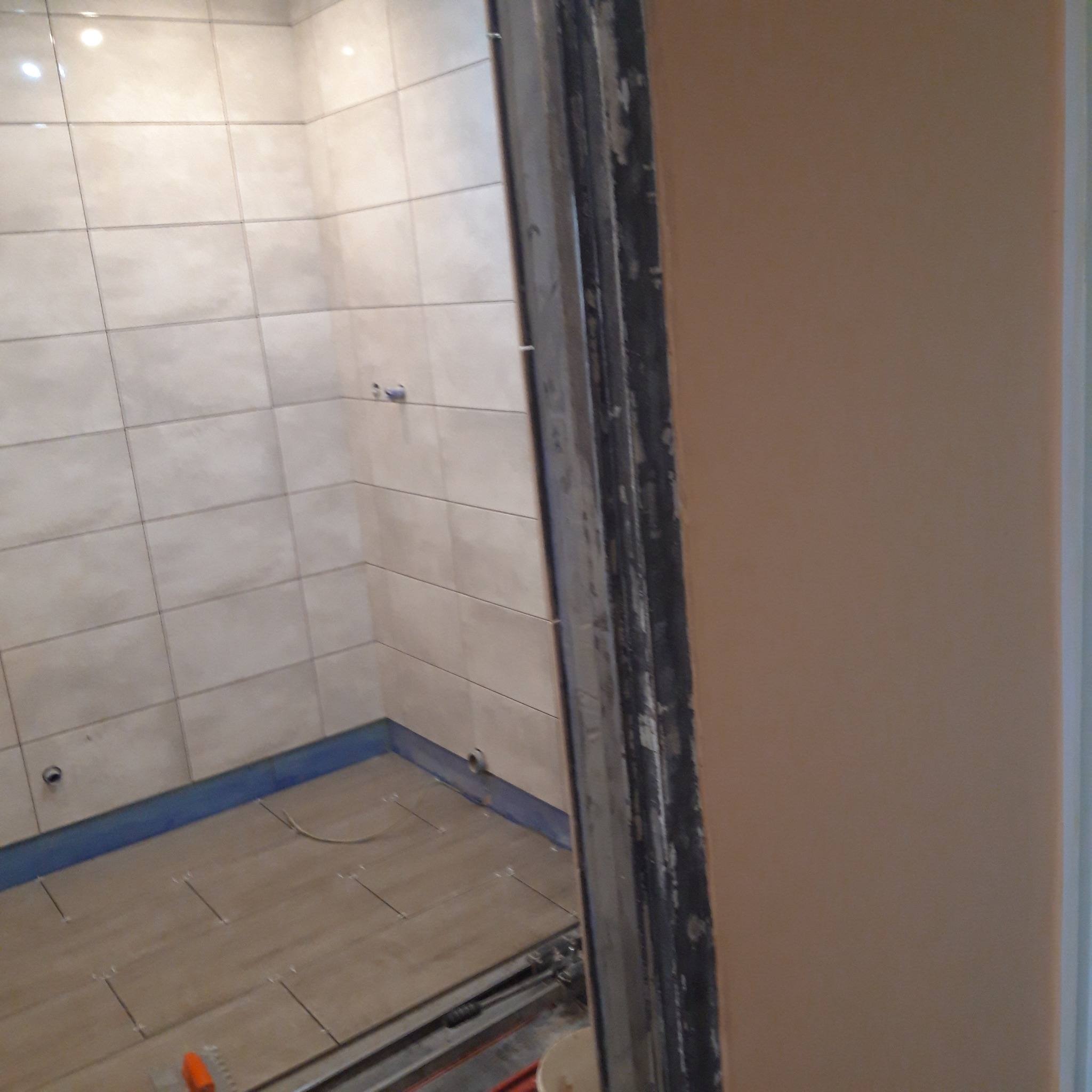 Wnętrze łazienki w trakcie układania płytek ceramicznych na podłodze i ścianach, widoczne krzyżyki dystansowe, narzędzie do cięcia płytek i niebieska hydroizolacja przy podłodze.