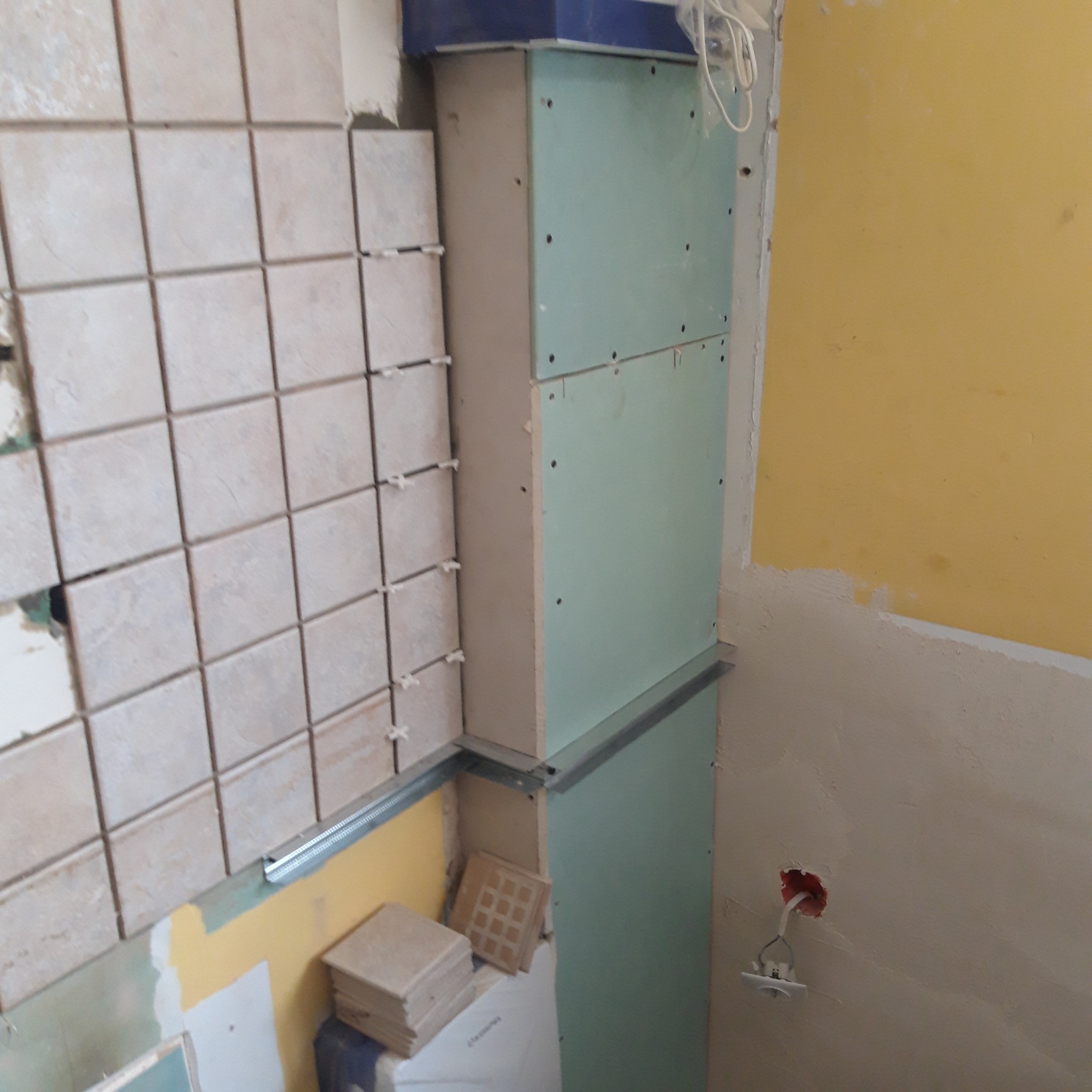 Wnętrze w trakcie prac: fragment ściany z położonymi płytkami ceramicznymi, obok konstrukcja z płyty kartonowo-gipsowej na metalowym stelażu, widoczne kable elektryczne.