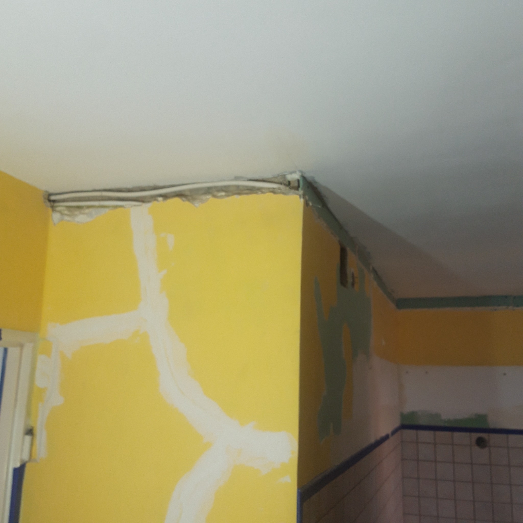 Żółta ściana z nałożoną białą gładzią w kształcie krzyża, widoczne także fragmenty instalacji elektrycznej pod sufitem i ściana z fragmentami zielonej farby oraz białe kafelki z ciemnoniebieską...