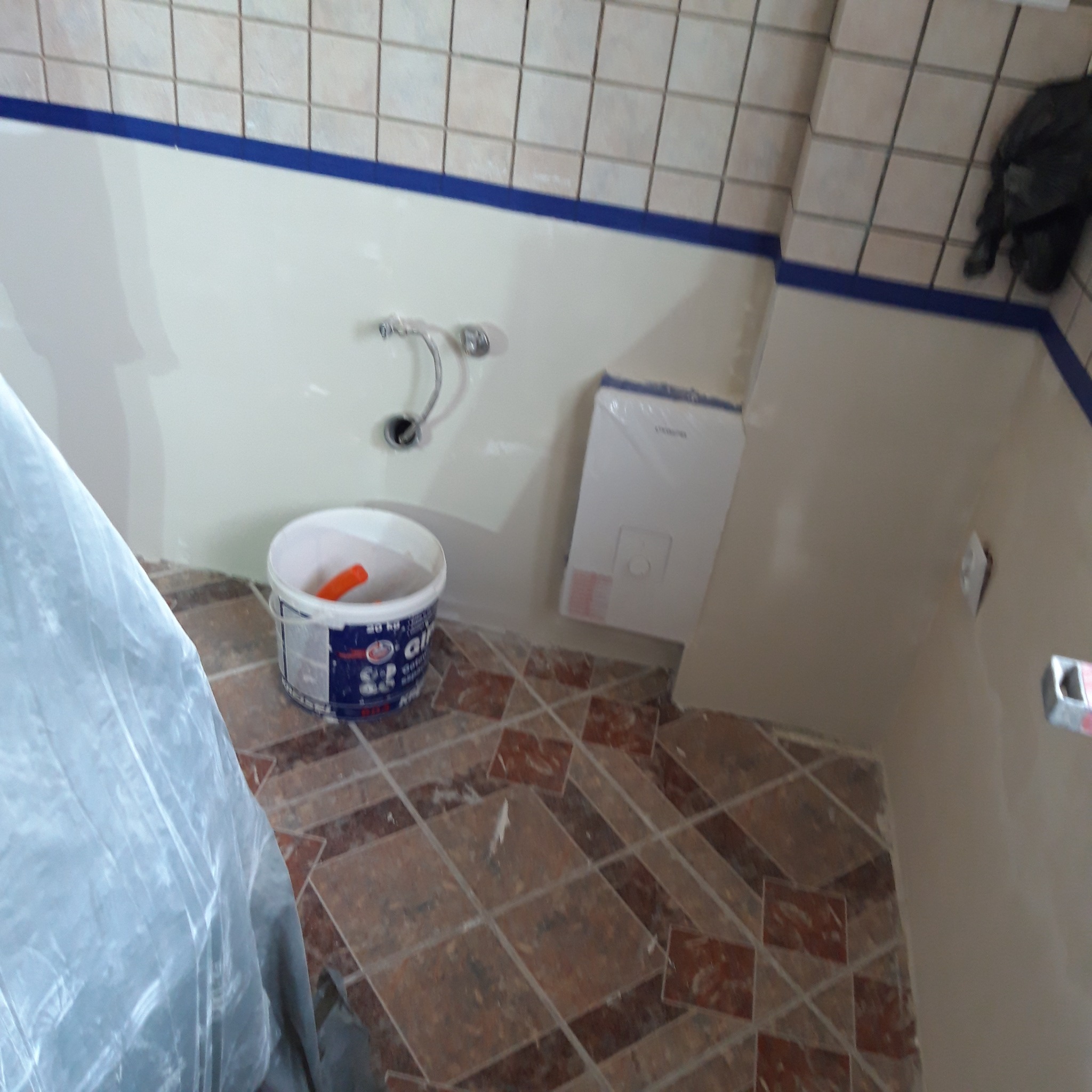 Przygotowania do malowania łazienki: wiadro z farbą, wałek malarski, folia ochronna na podłodze z płytek w odcieniach brązu, świeżo pomalowane ściany w jasnym kolorze z niebieską taśmą malarską...