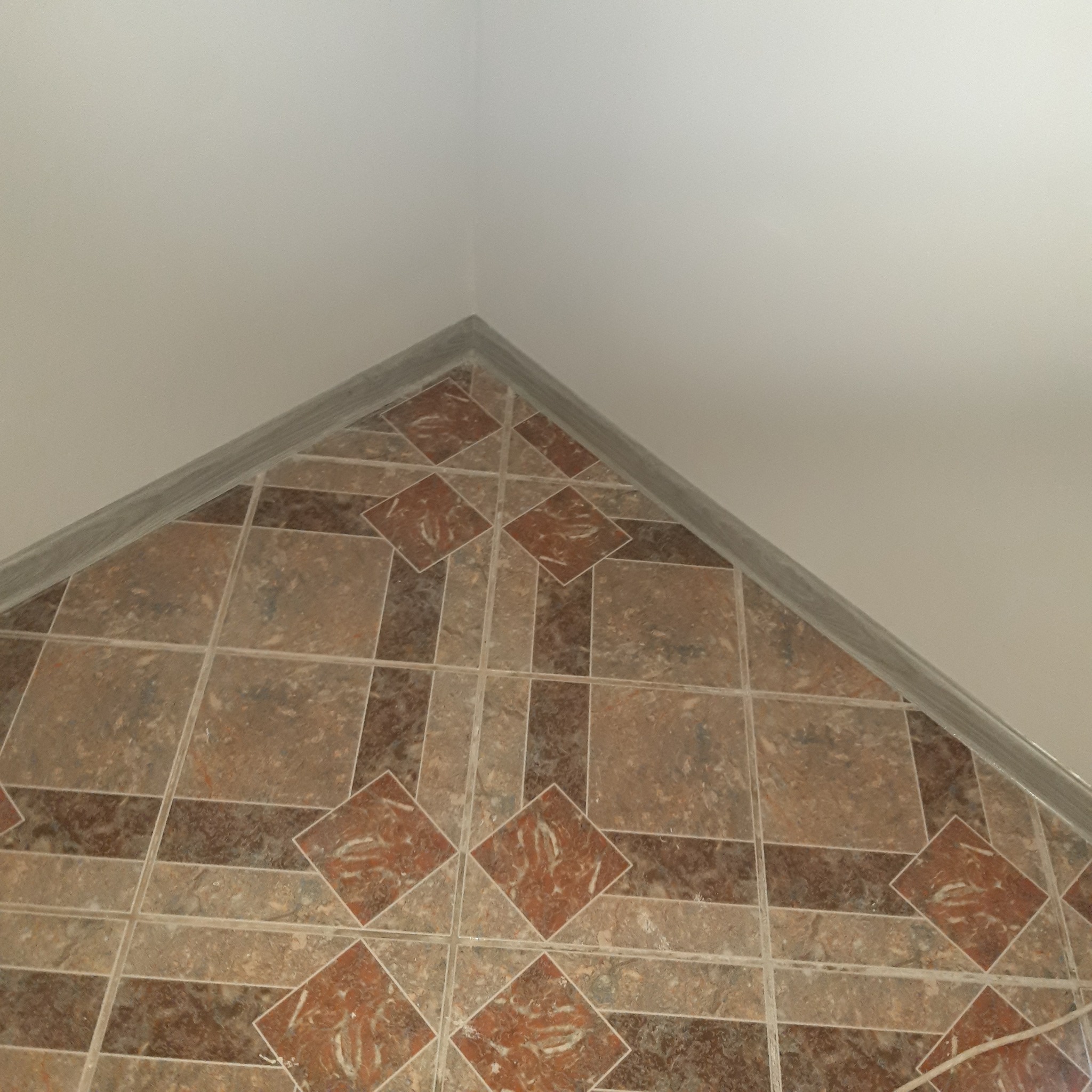 Ukończona podłoga z płytek ceramicznych w odcieniach brązu i beżu, ułożonych w geometryczny wzór z rombami, widoczna przy krawędzi ściany i sufitu.