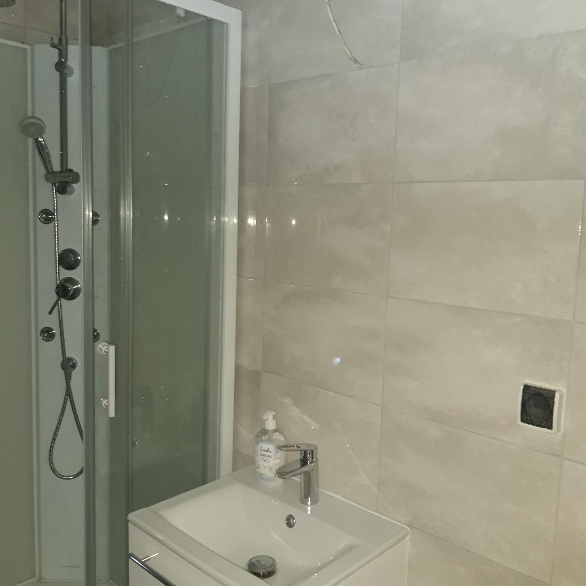 Wykończona łazienka z kabiną prysznicową z hydromasażem, umywalką z szafką i baterią oraz beżowymi płytkami na ścianach; widoczny dozownik mydła i gniazdko elektryczne.