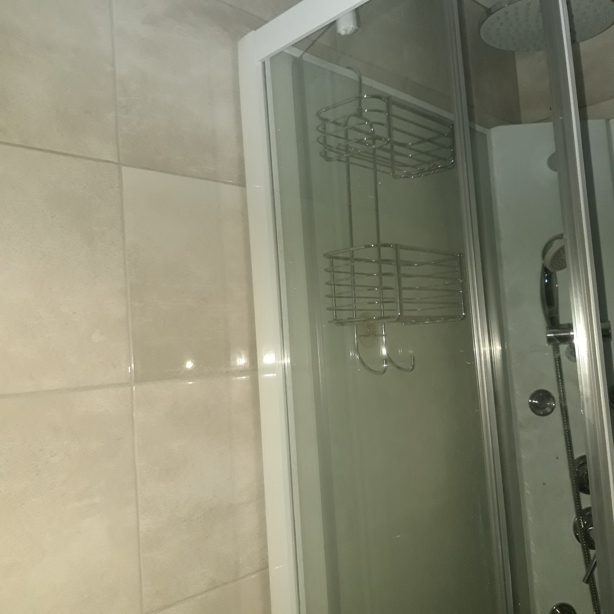 Szklana kabina prysznicowa z metalową półką na kosmetyki, widoczne beżowe płytki na ścianie.